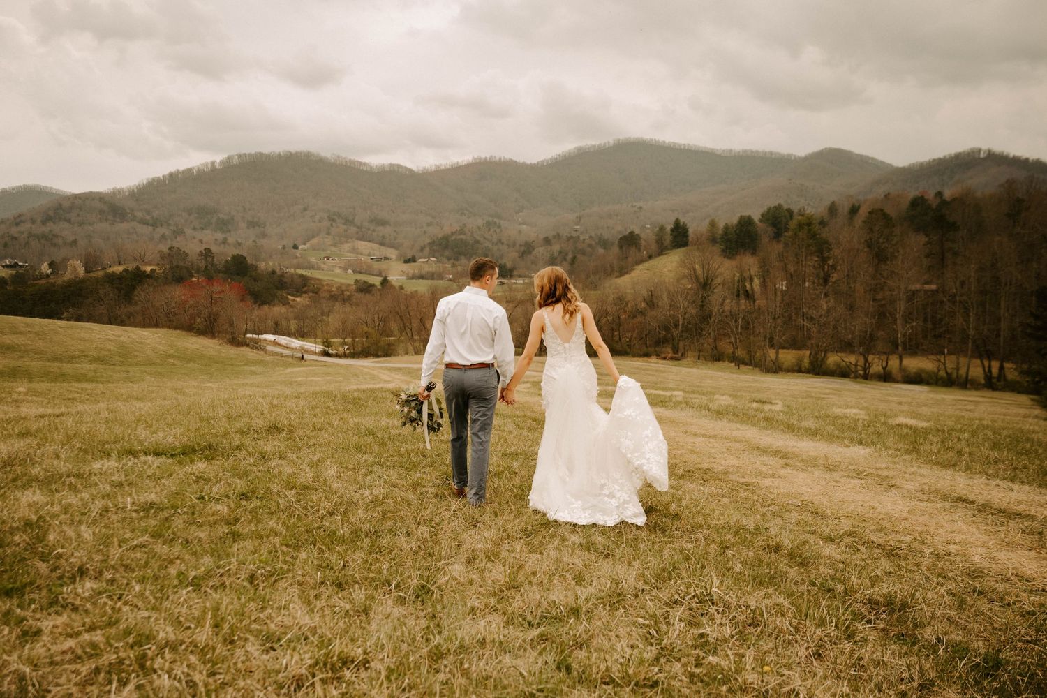 A couple walks hand in hand across a grassy meadow with rolling mountains in the distance on an overcast day.
