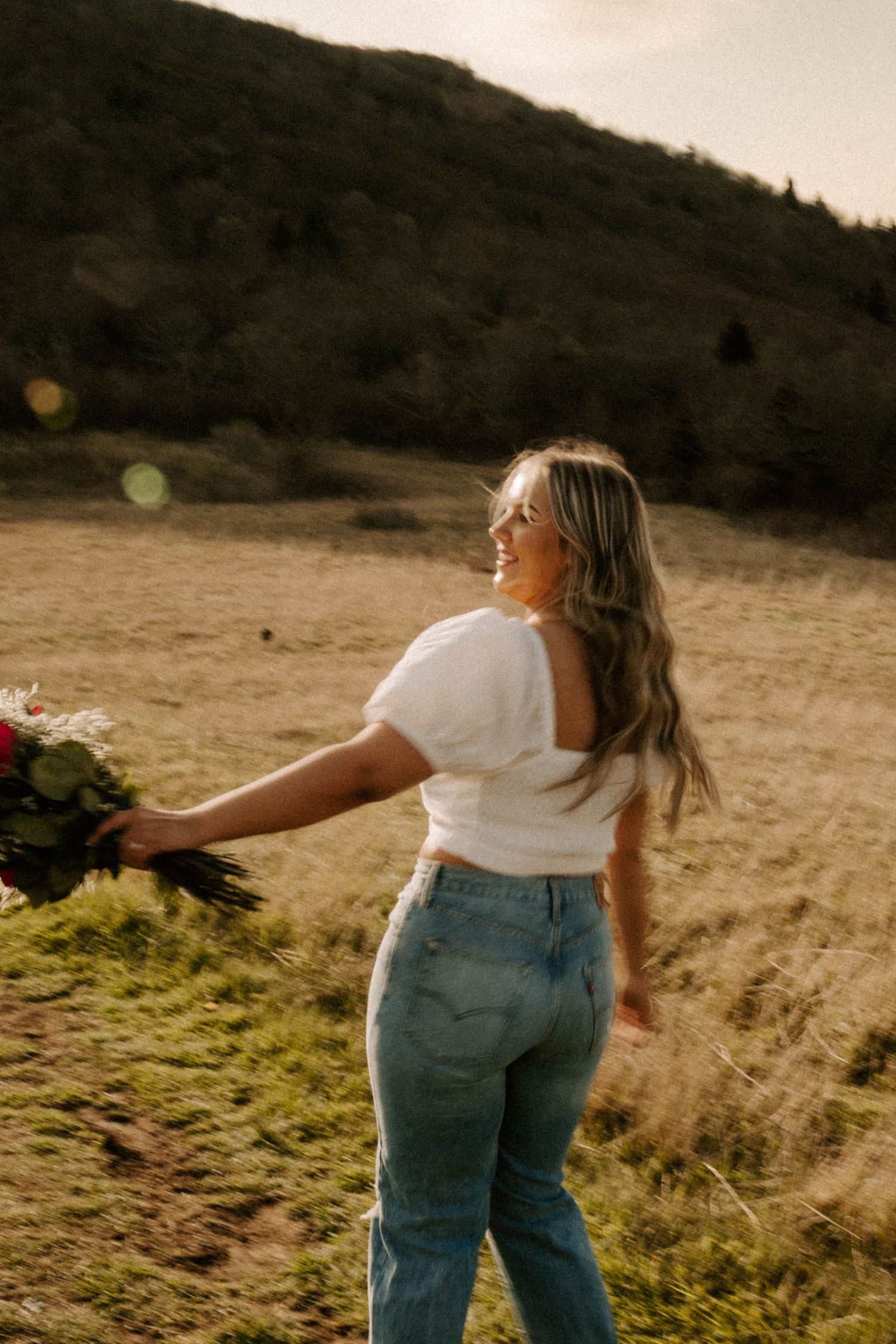 A person in a white top and jeans holding flowers stands in a golden field at sunset with hills in the background.