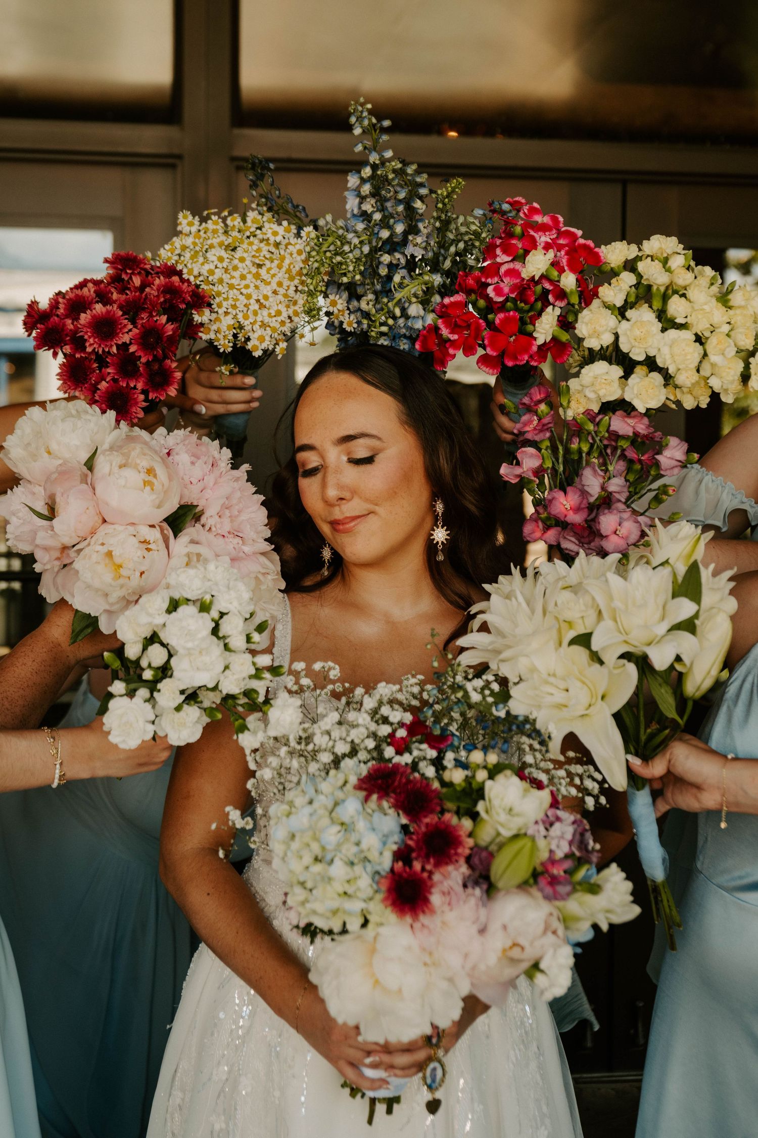 A bride in a white dress stands surrounded by bouquets of red, pink, and white flowers with baby's breath accents.