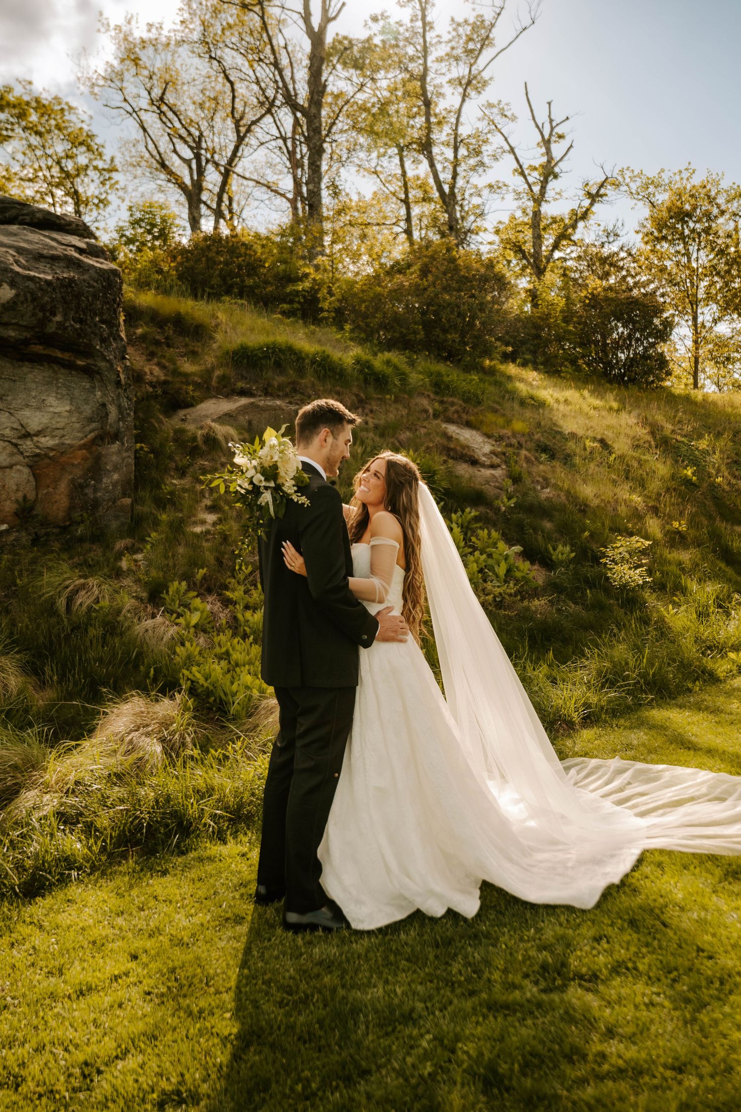 A romantic wedding couple embraces on a grassy hilltop at sunset with a flowing white veil against natural rock formations.