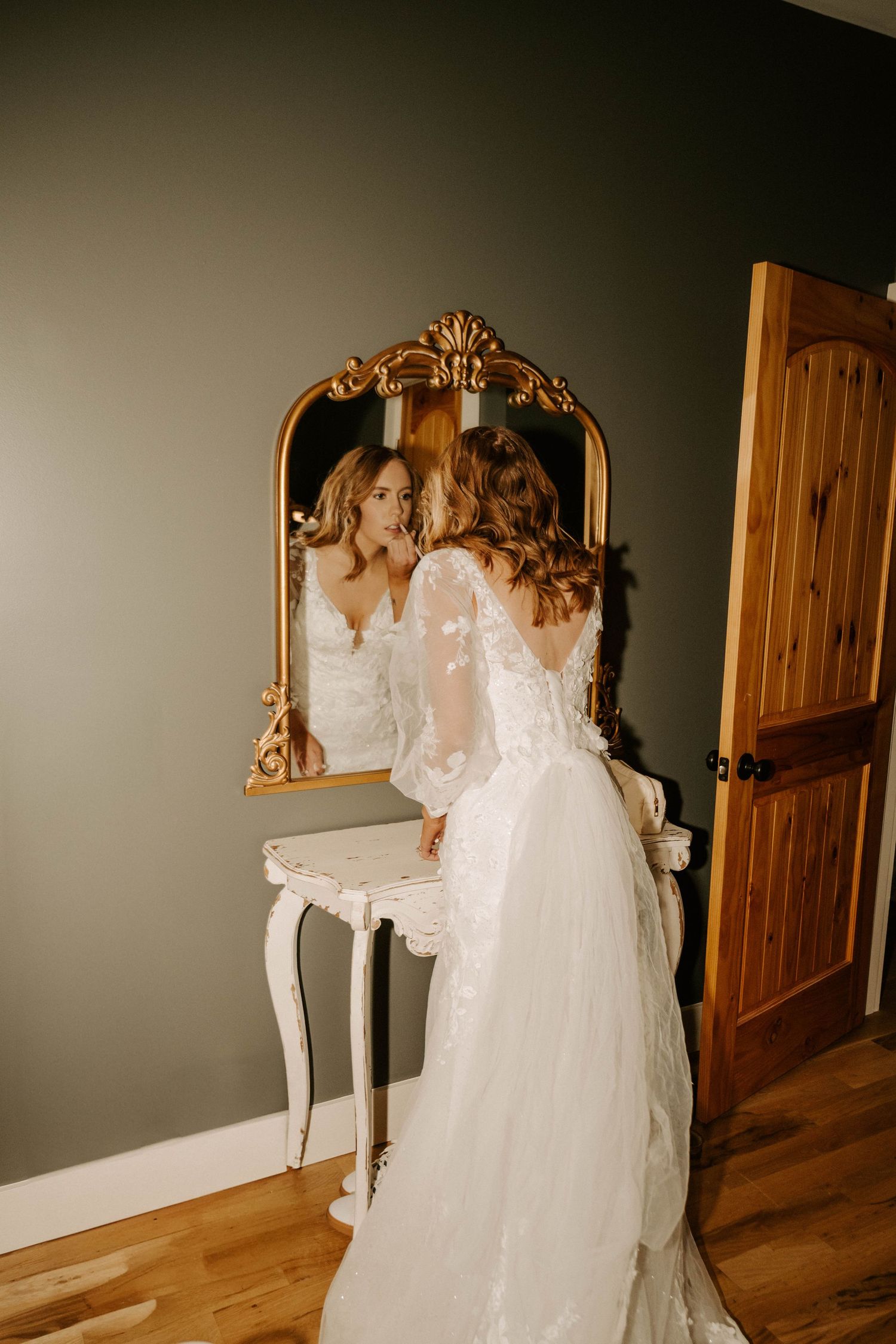 A vintage-style vanity with ornate gold mirror reflects the back of someone in a flowing lace wedding dress.