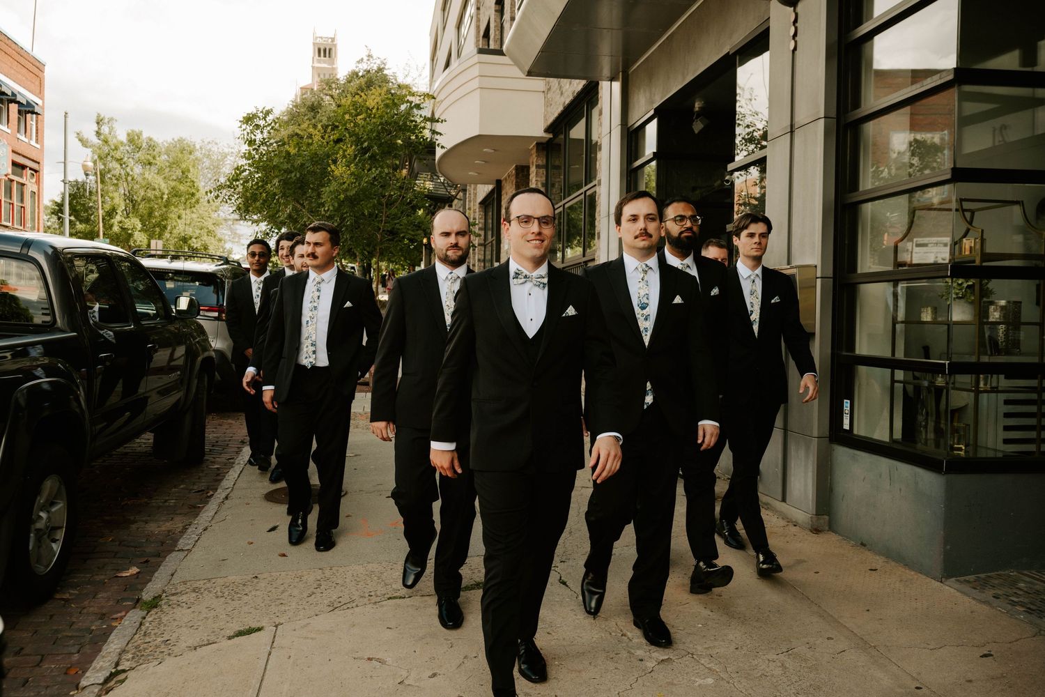 A group in formal black tuxedos walk together down a city sidewalk on a sunny day.