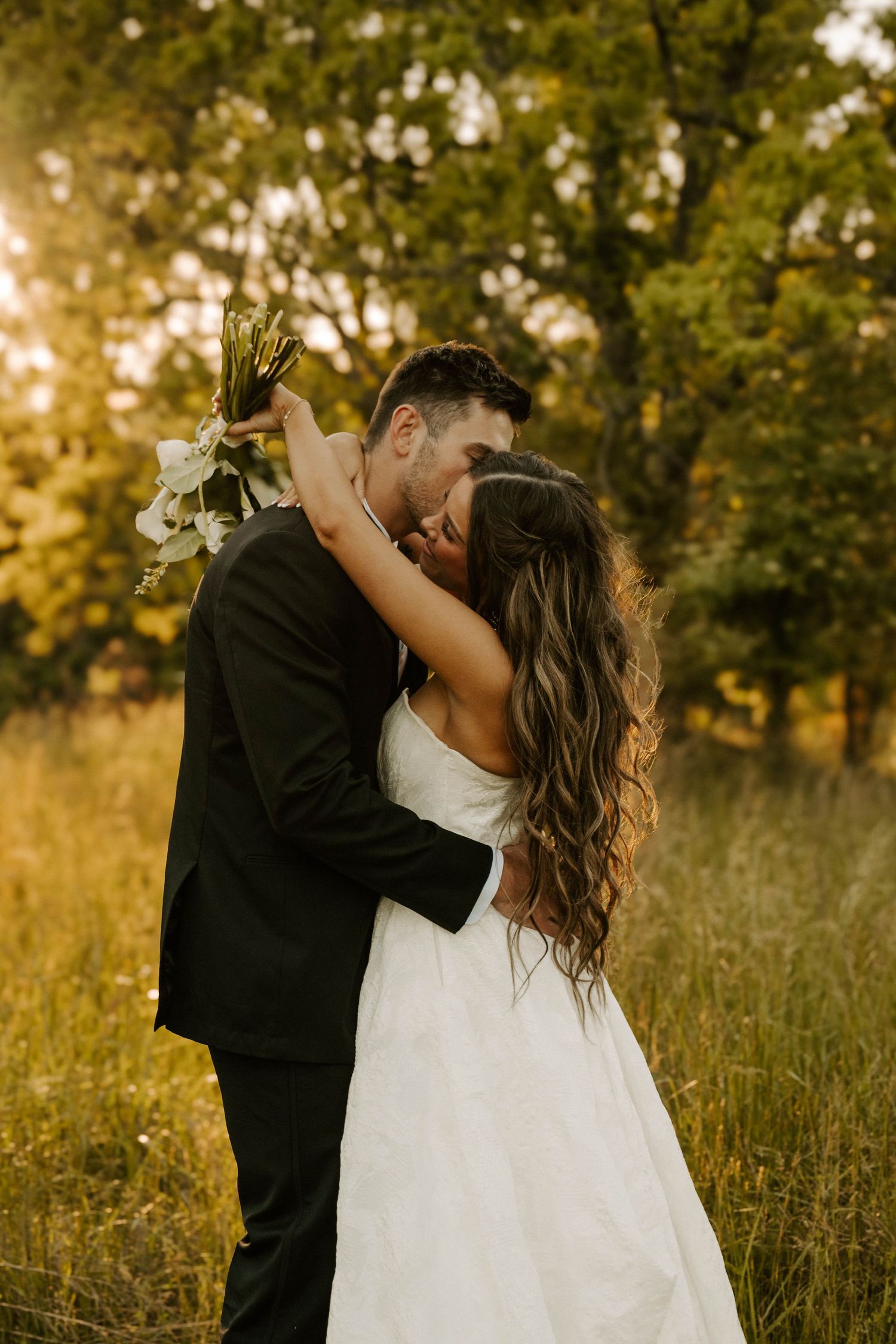 Couple sharing romantic embrace in golden sunlit meadow with tall grass and trees during sunset wedding photoshoot.