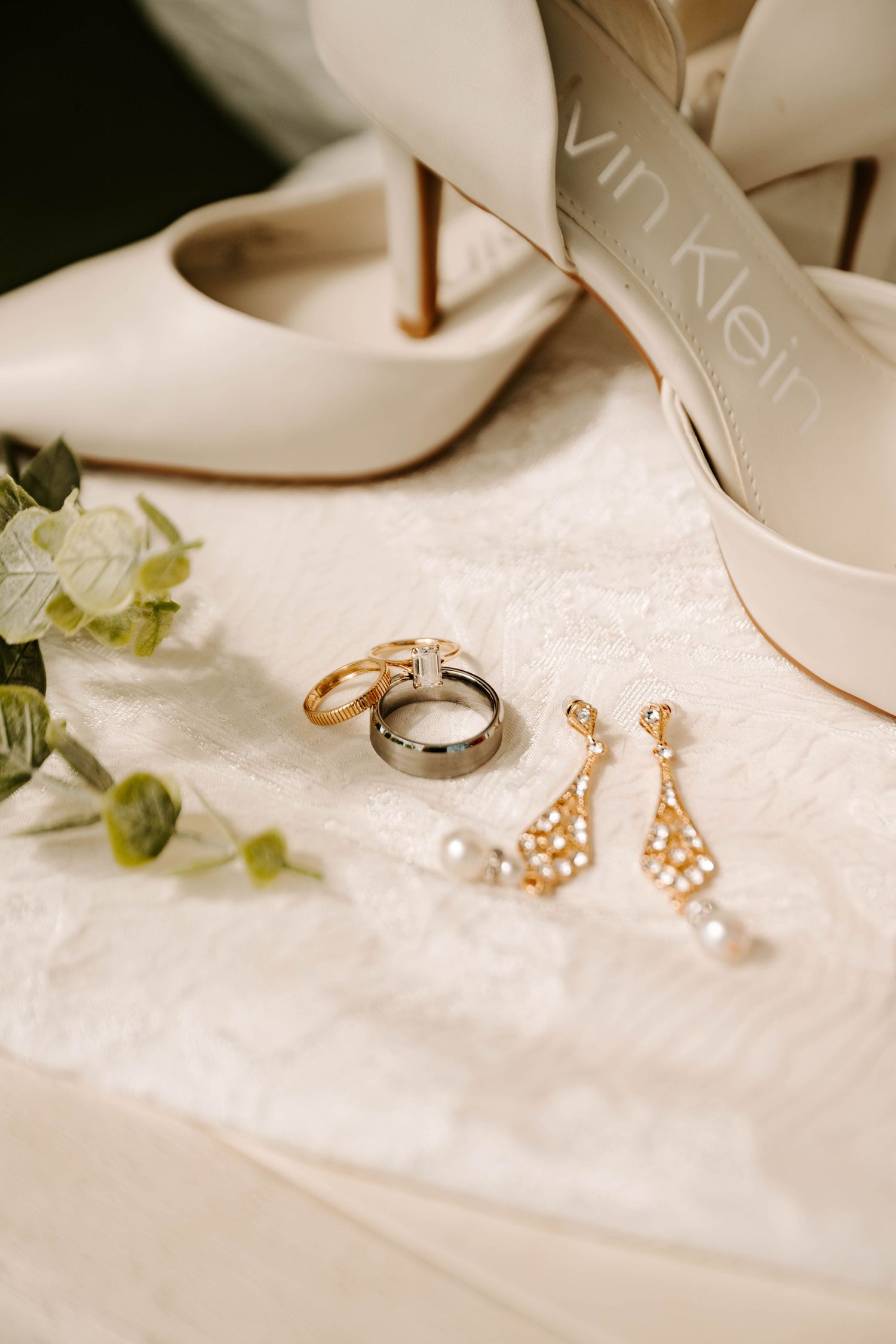 Wedding accessories including white heels, gold dangle earrings, wedding rings and delicate green florals on white fabric.