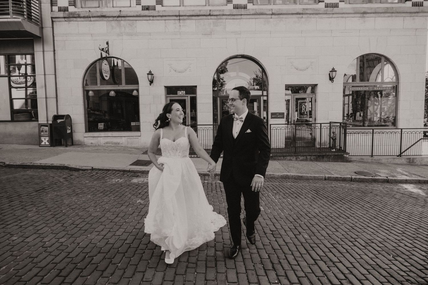 A couple in formal wedding attire walks along a cobblestone street in front of an elegant building with arched windows.