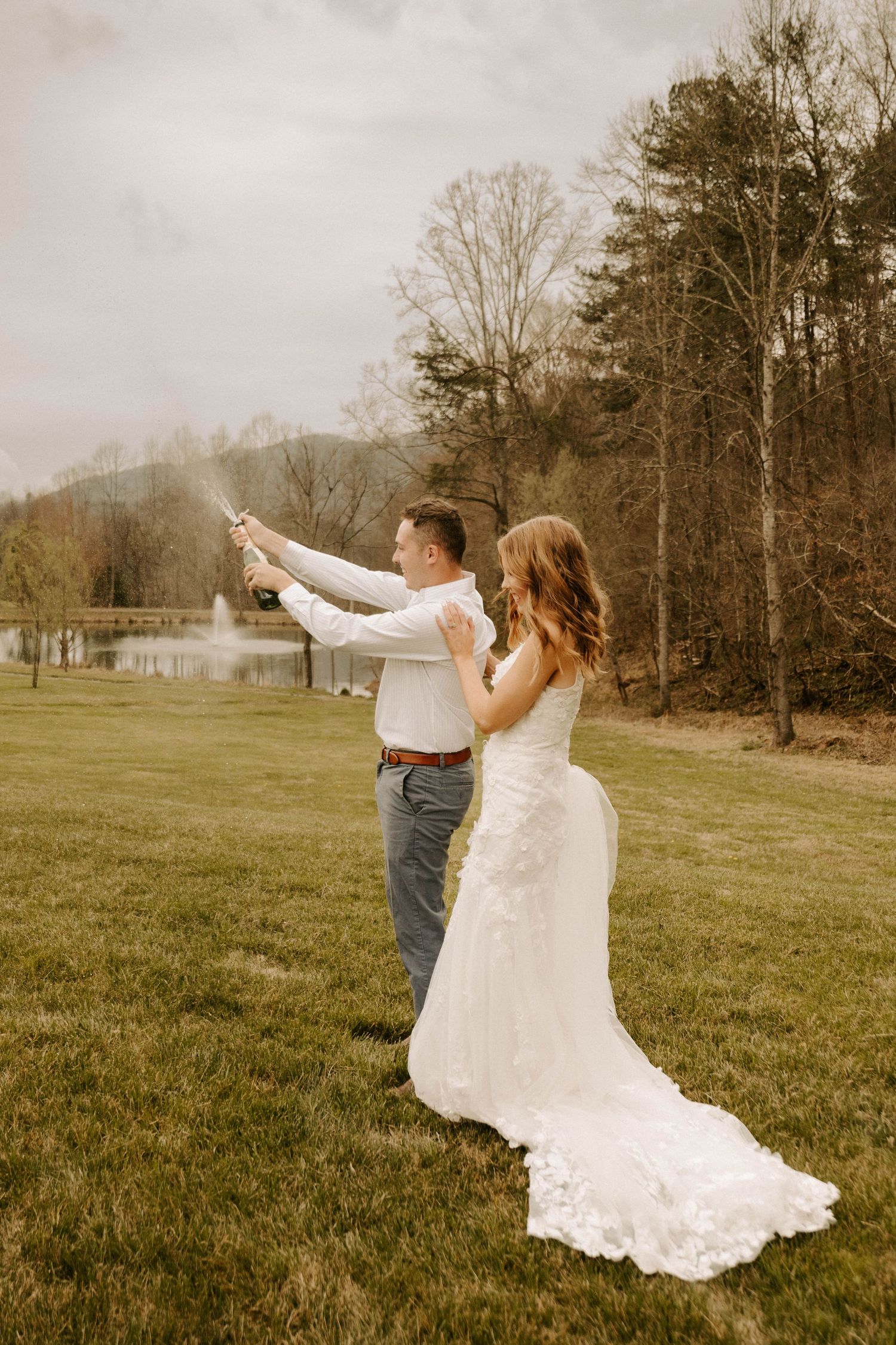 A couple in wedding attire stands on a grassy hillside pointing at something in the distance on an overcast day.