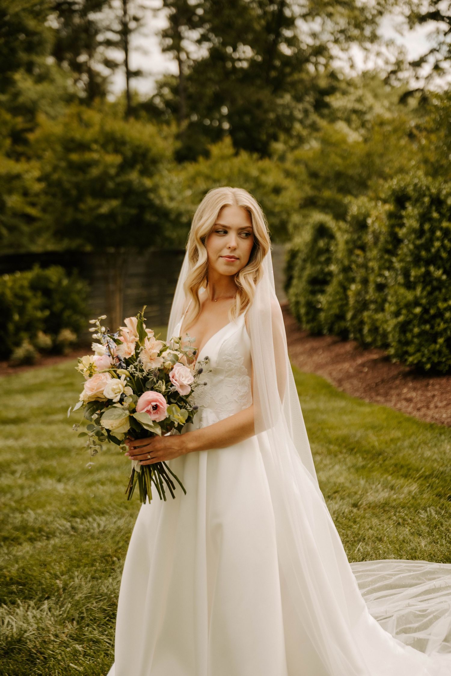 A bride in a flowing white dress holds a beautiful bouquet of pink and white flowers in a garden setting.
