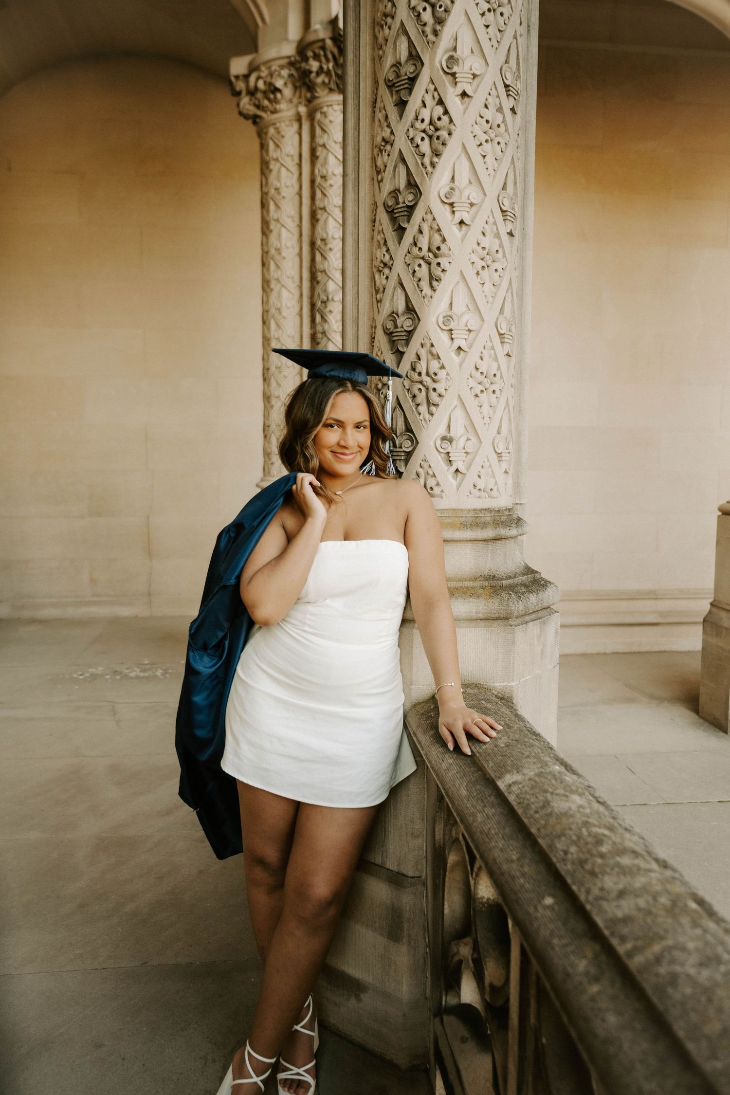 Graduate in blue cap and gown with white dress poses against ornate stone architecture in warm lighting.