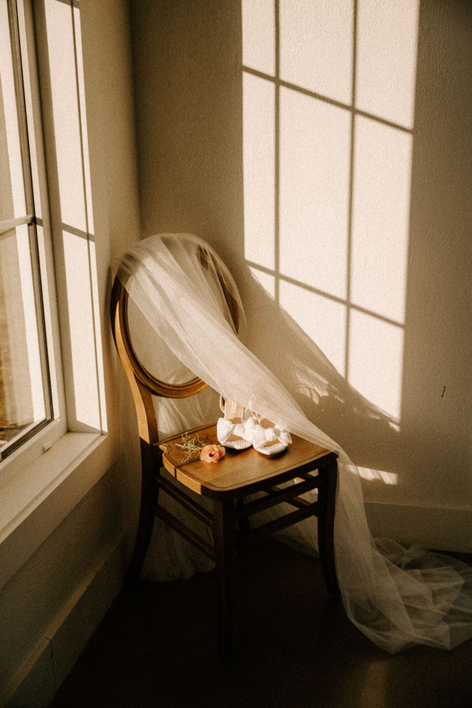 A flowing white wedding veil drapes over a wooden chair by a sunlit window with wedding shoes placed on the seat.