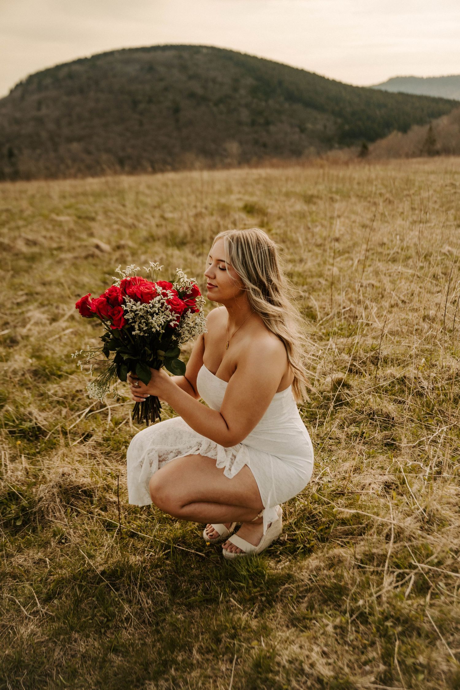 A person in a white dress crouches in a grassy field holding a bouquet of red roses against a mountainous backdrop.