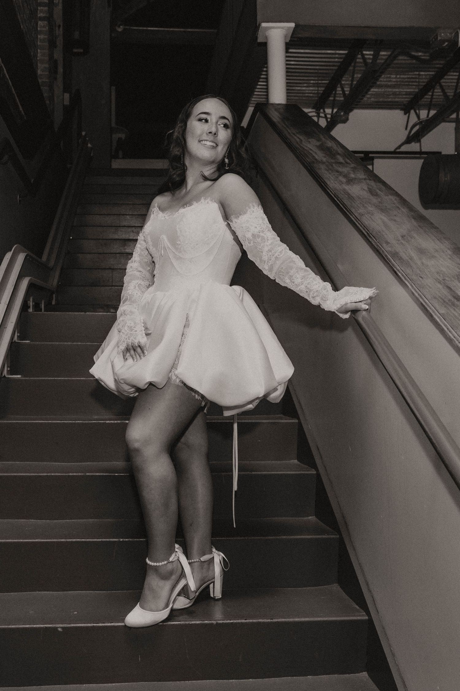 A dancer poses on stairs wearing a white long-sleeved mini dress and heels in a black and white vintage-style photo.
