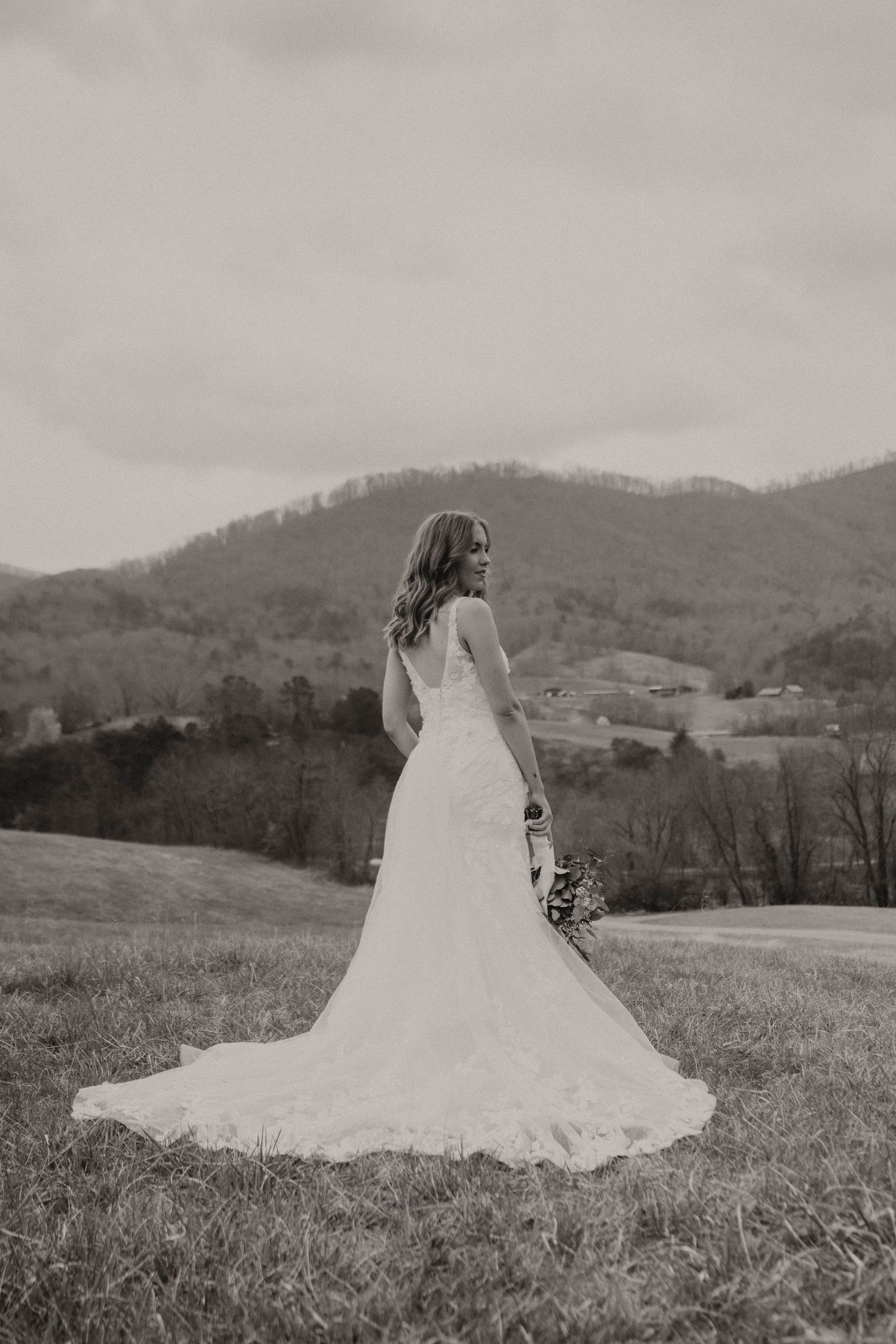 Black and white photo of bride in lace wedding dress with flowing train standing on hillside with mountain landscape view.