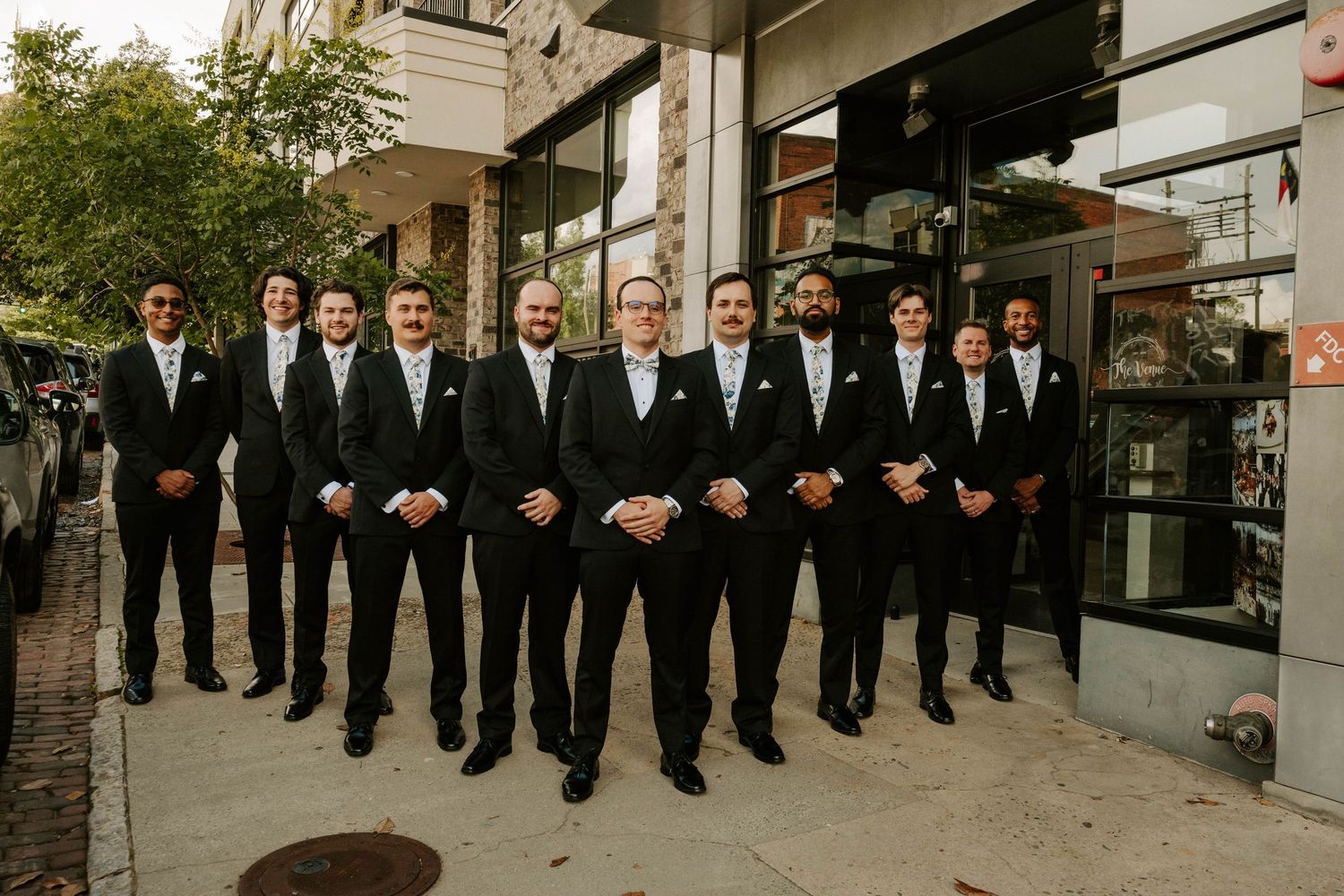 Wedding party in black suits poses outdoors against a modern building facade in natural light.