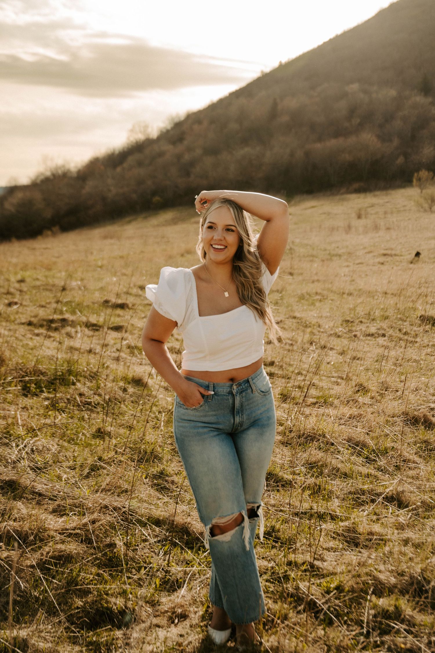 A person in ripped jeans and white crop top poses in a golden field at sunset with mountains in the background.