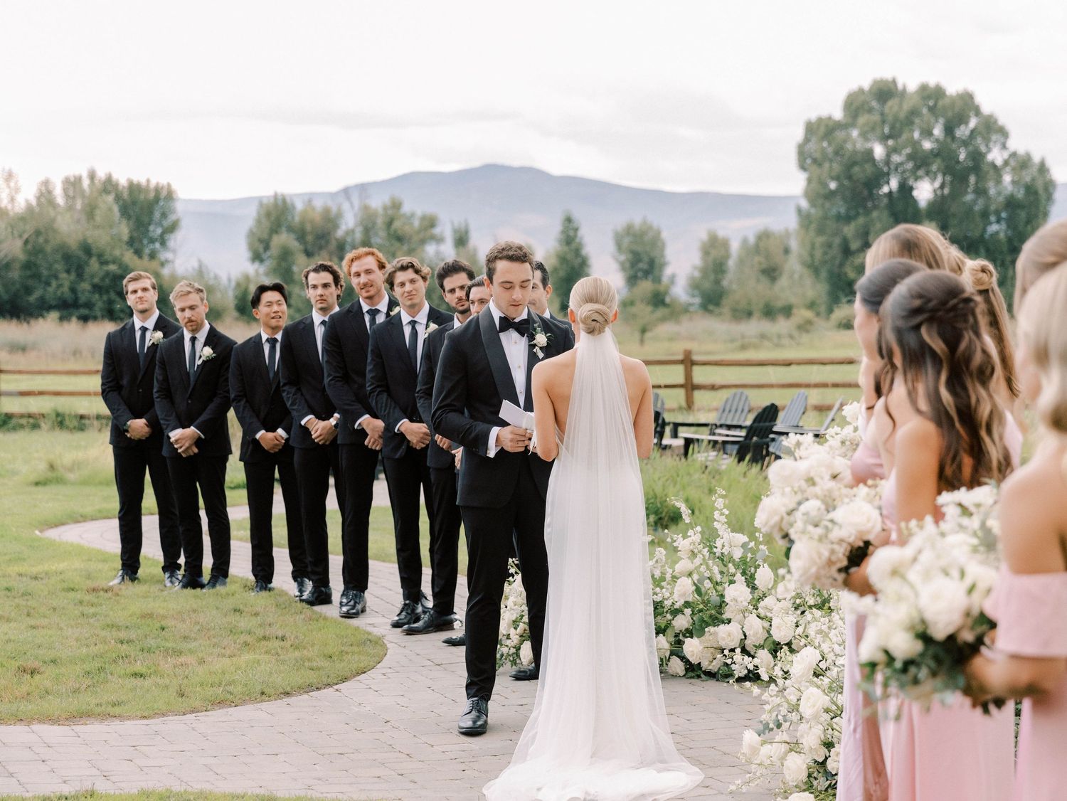 Groomsmen in black suits line up during outdoor mountain wedding ceremony with floral decor.