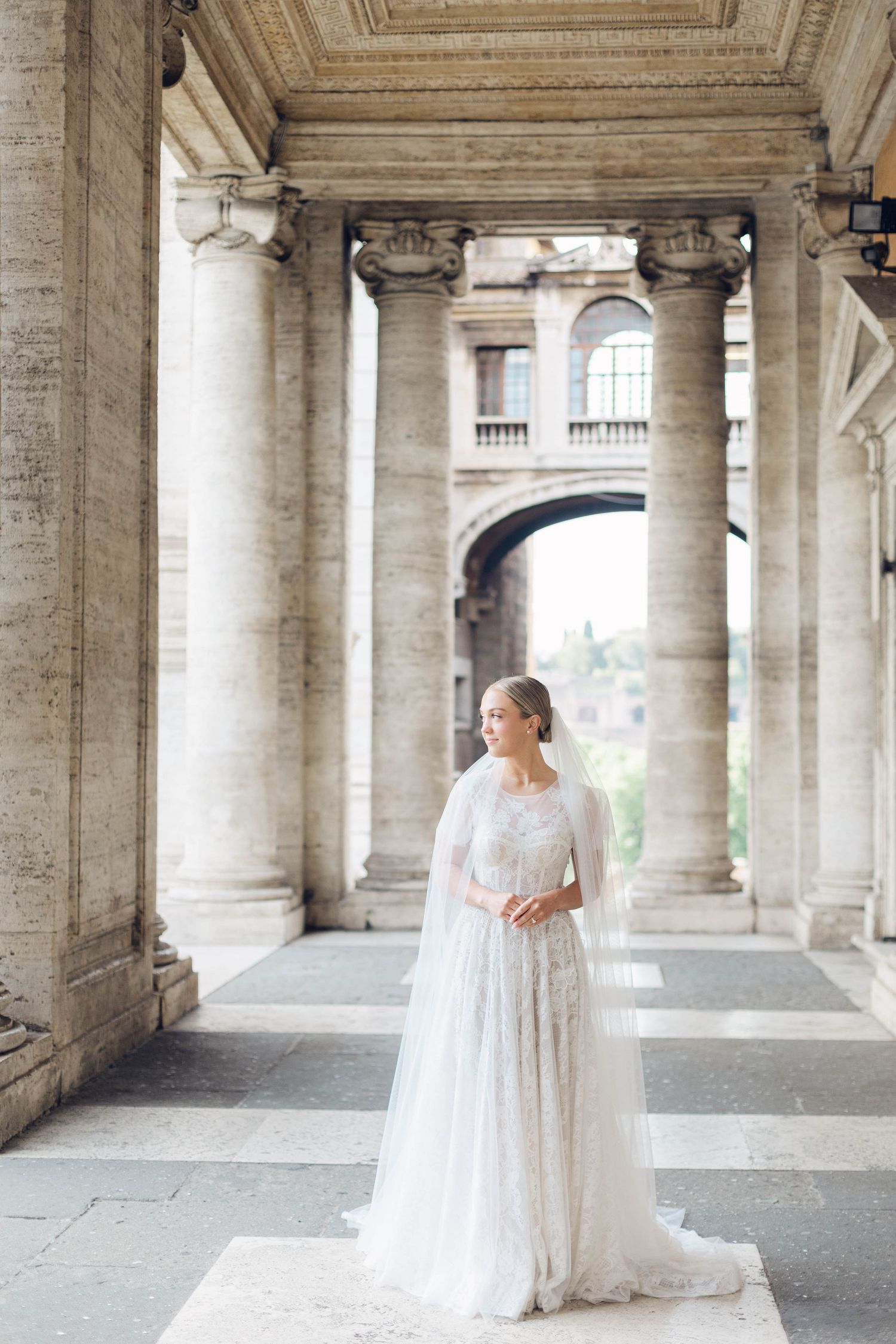 A bride in a lace white gown stands alone in a classical stone colonnade of rome italy.