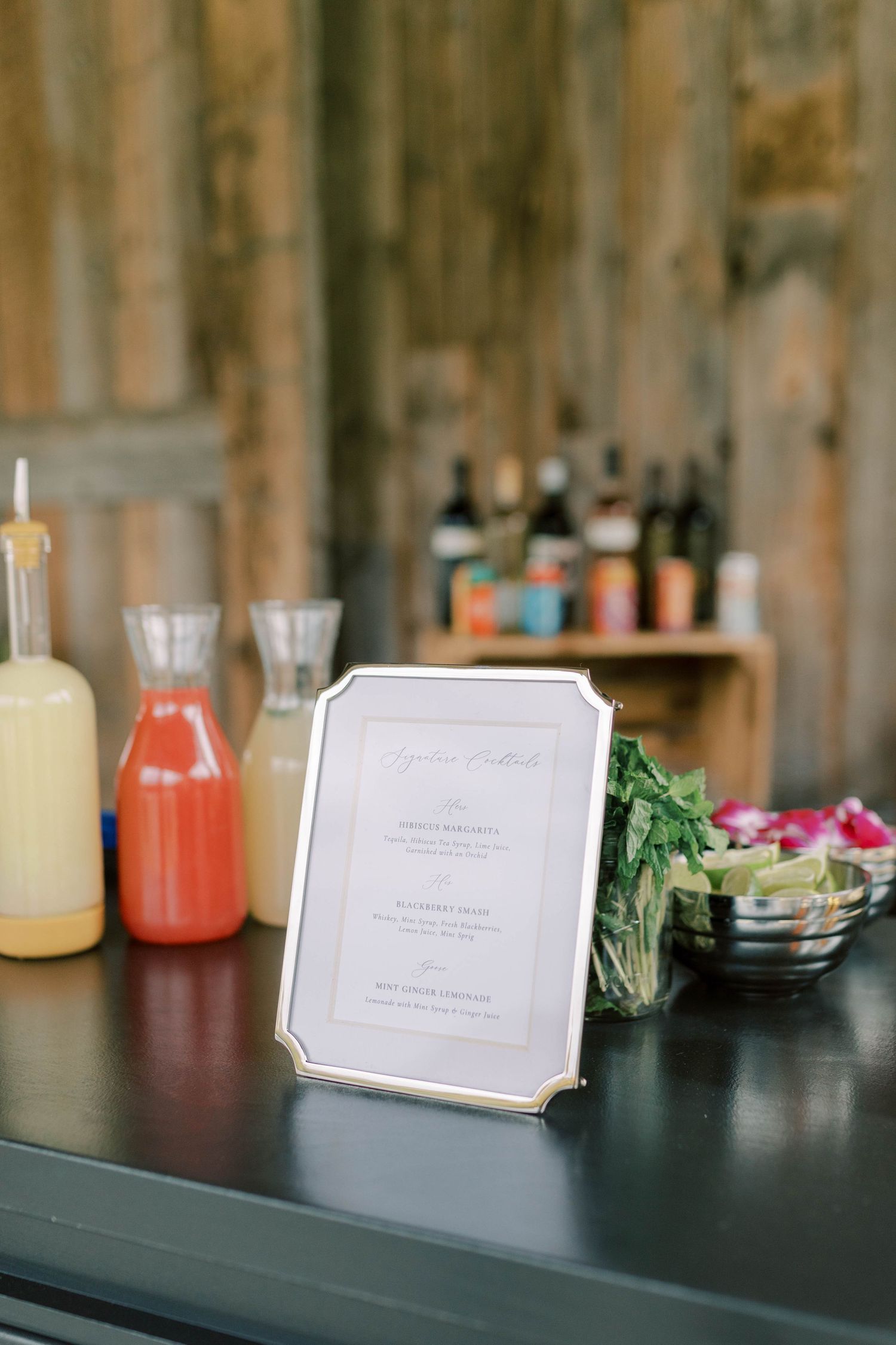 Wedding bar setup with drink dispensers and framed menu on rustic wooden backdrop.