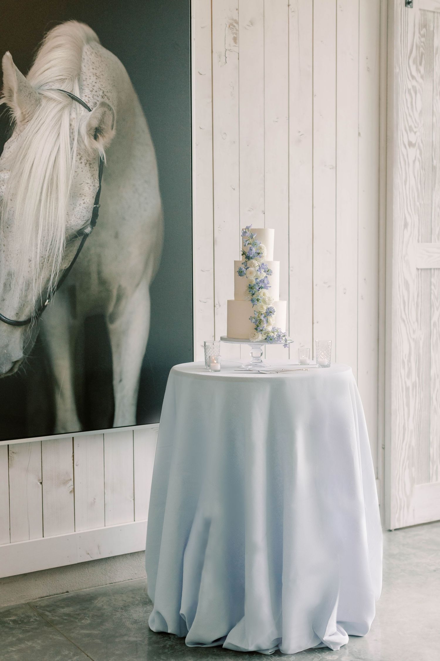 White wedding cake displayed on draped table beside large horse portrait in rustic setting.