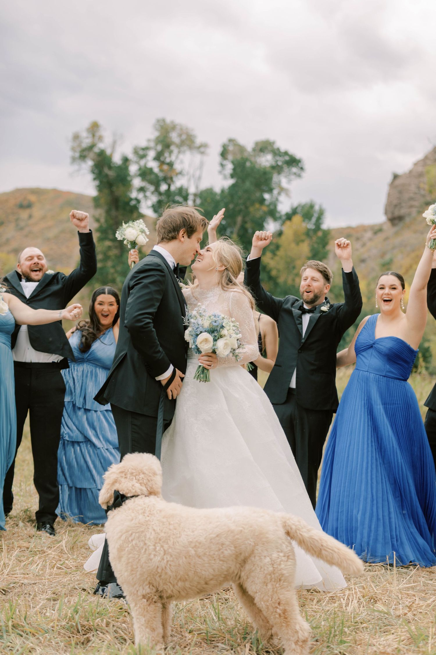 Wedding party celebrates with arms raised while fluffy dog photobombs their joyful moment.