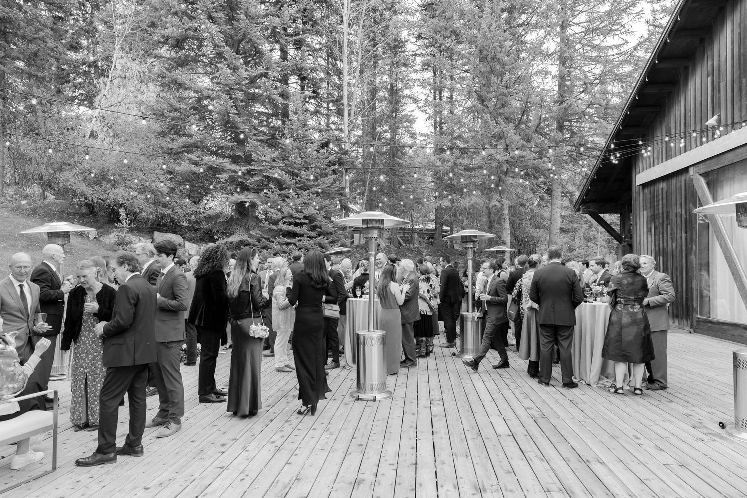 Vintage black and white photo of guests gathering on a wooden deck surrounded by forest.