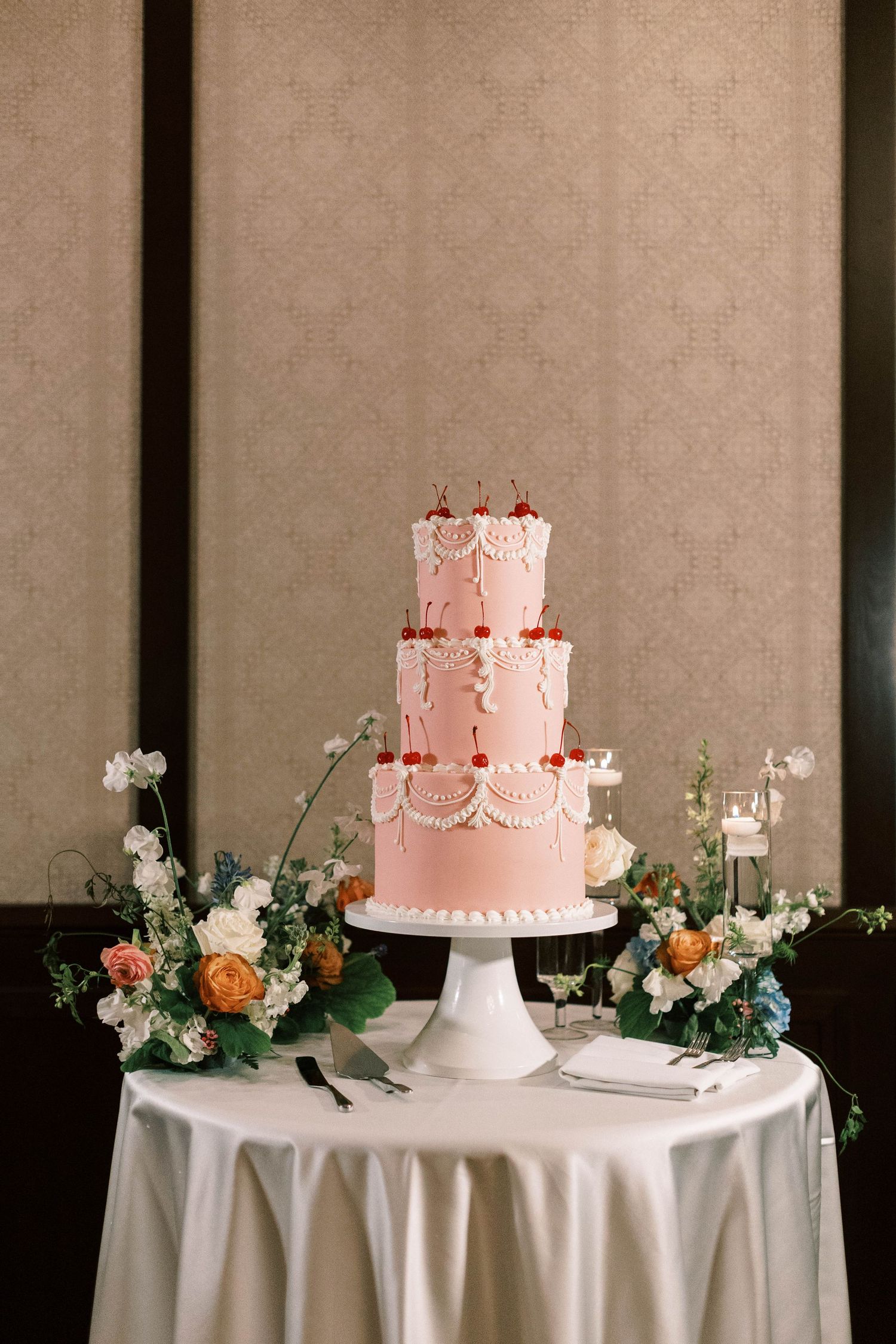Three-tiered pink wedding cake with delicate floral decorations displayed on a white table with fresh flower arrangements.