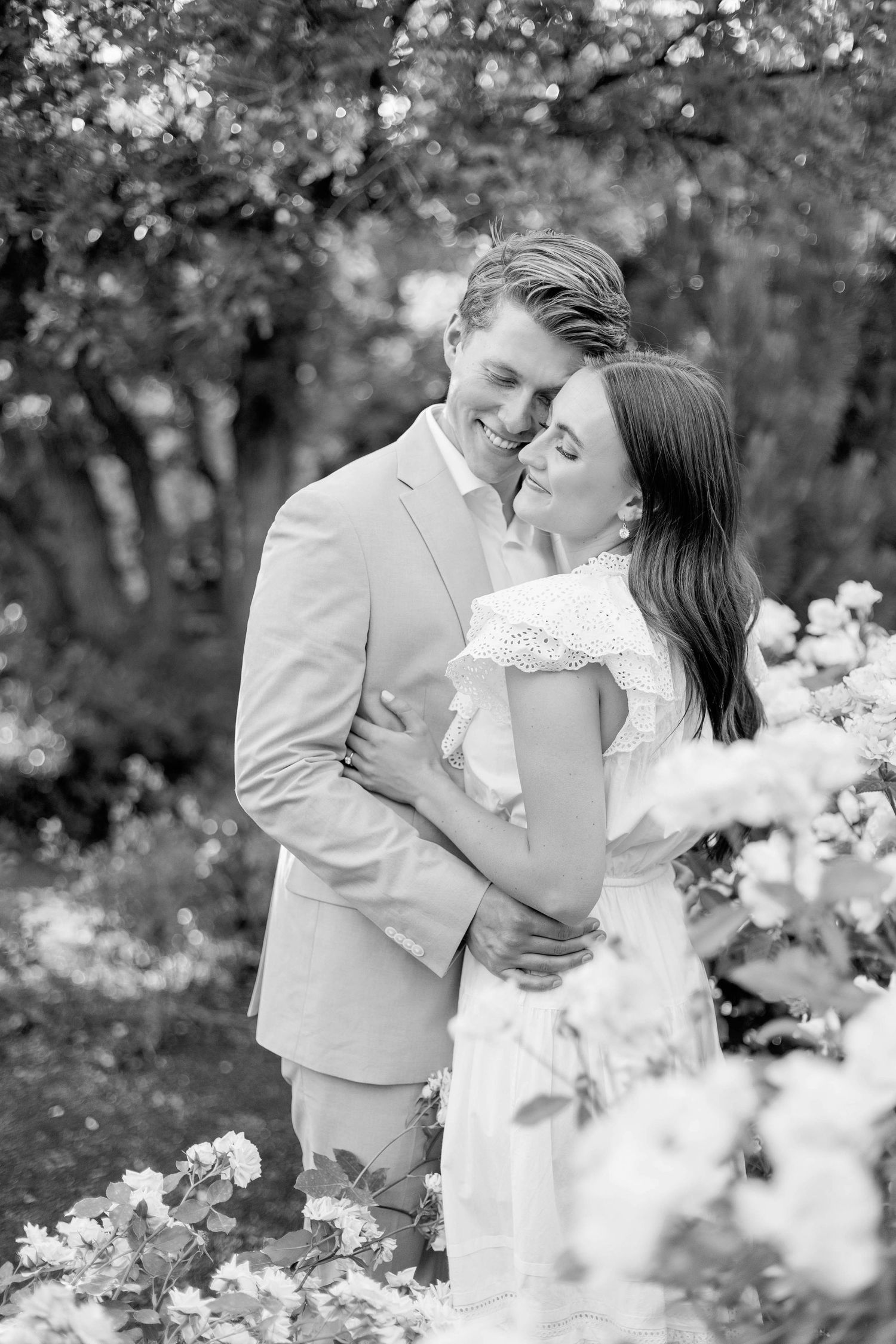 A black and white portrait of a couple sharing a tender moment among blooming flowers.