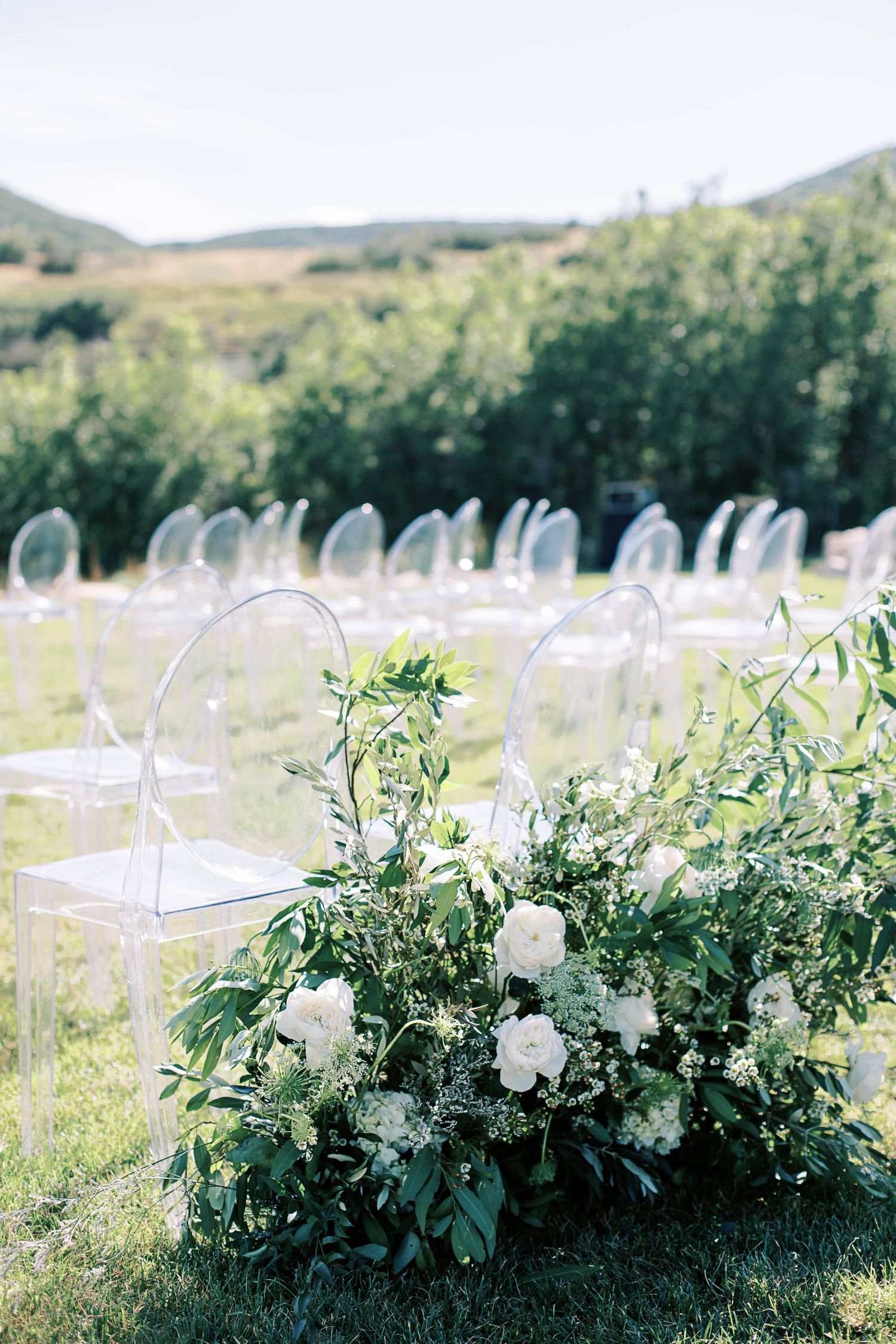 Ghost chairs and lush white floral arrangements set up for outdoor mountain wedding ceremony.