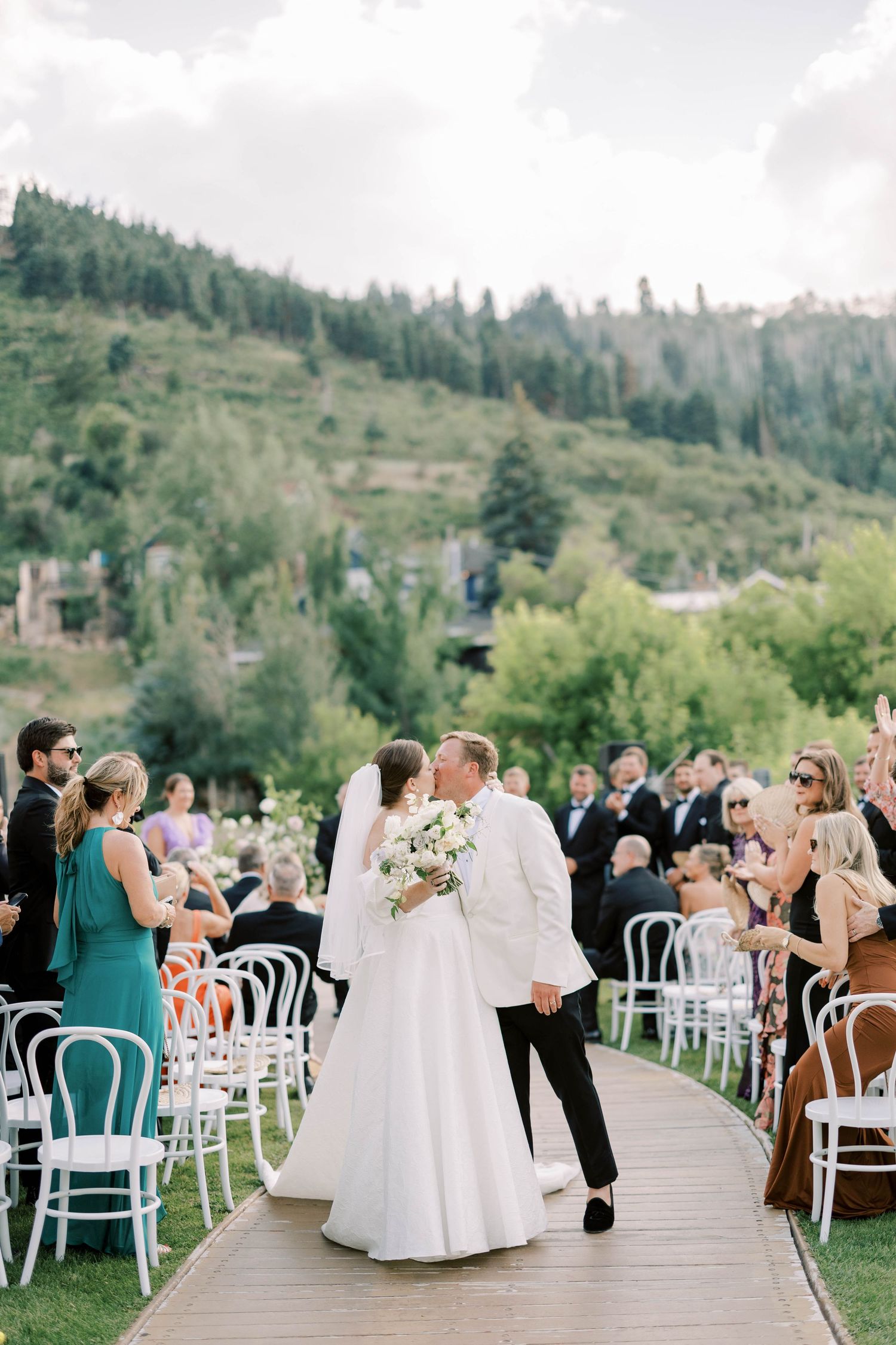Newlyweds walking down aisle at mountain resort wedding with seated guests and white chairs.