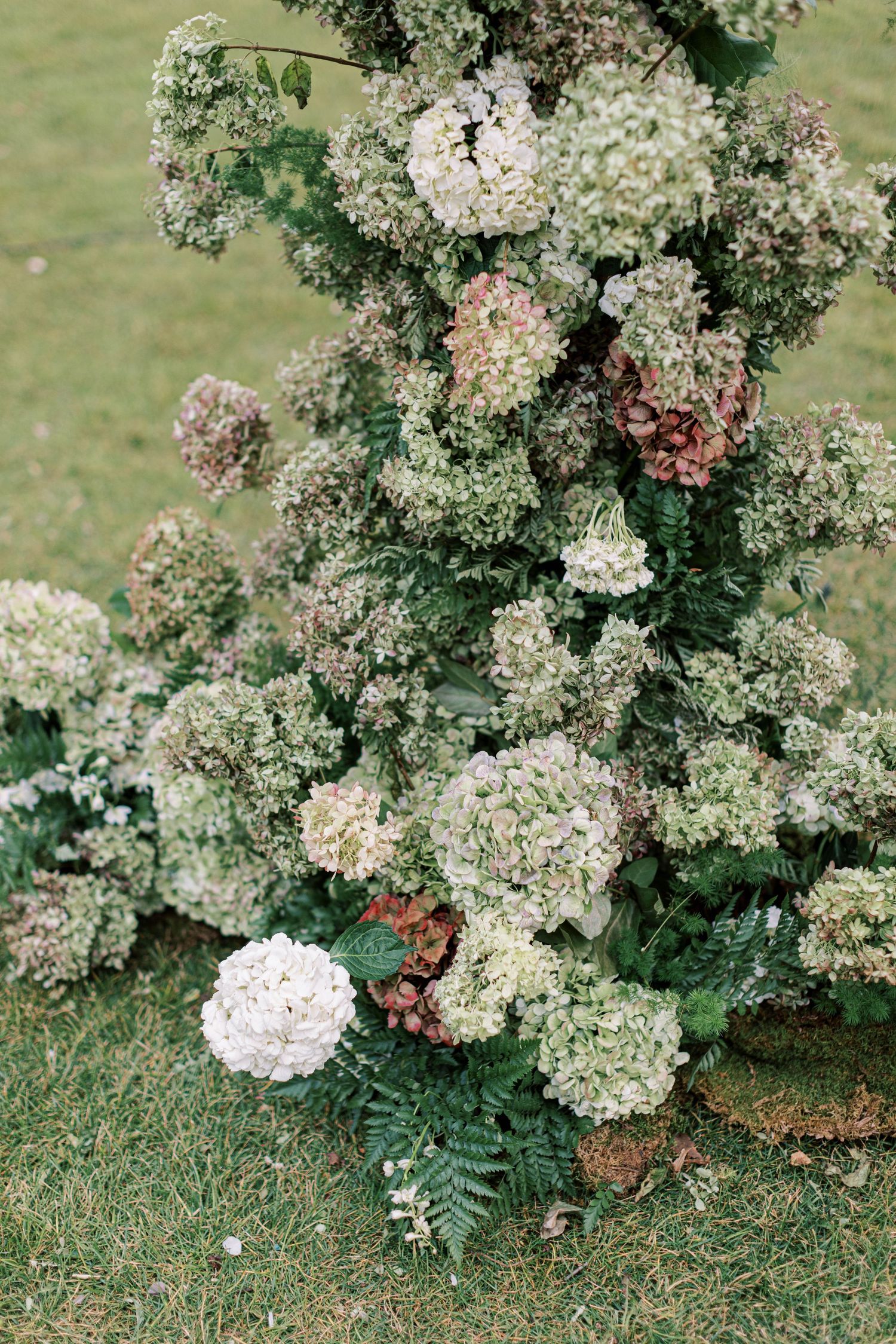 Lush floral arrangement of white and blush hydrangeas with greenery on grass.
