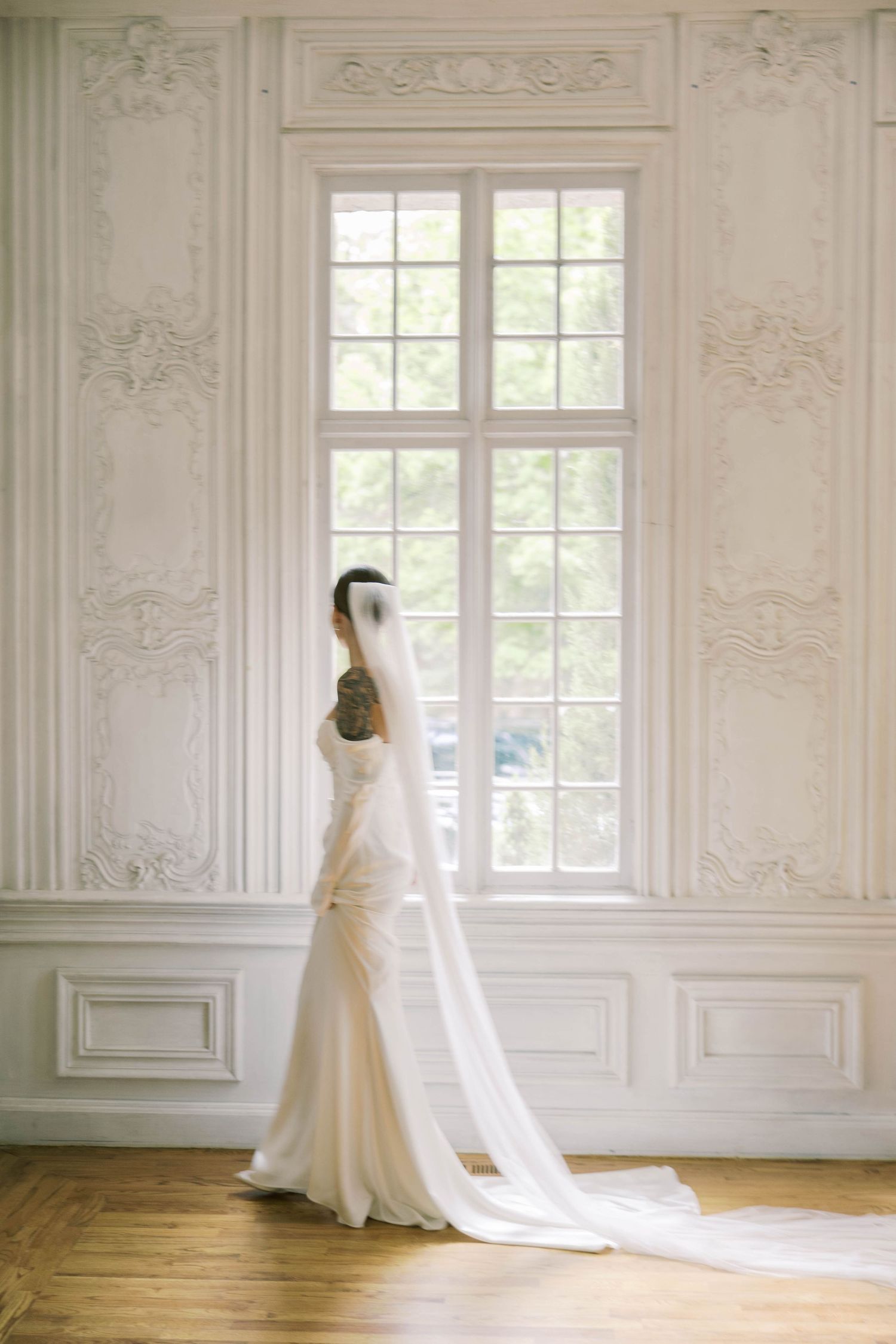 Elegant white wedding gown with flowing train photographed against ornate white paneled walls and tall windows.