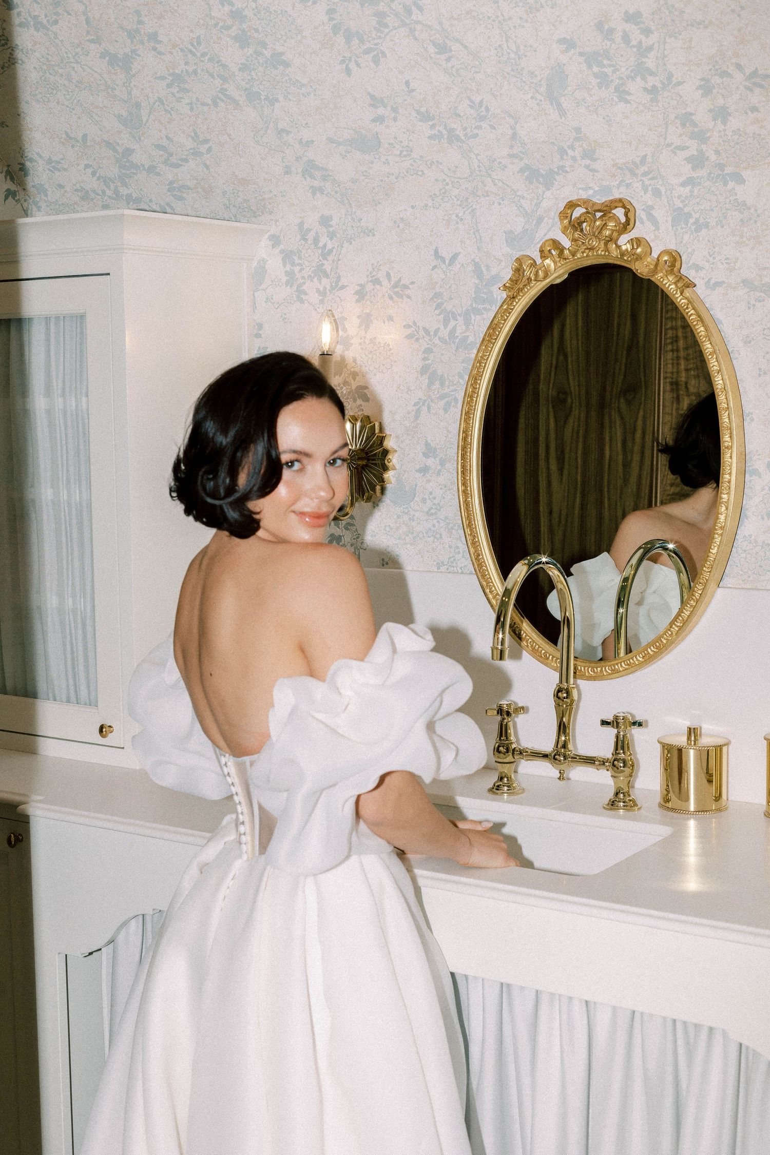 Elegant bride in luxurious white off-shoulder gown poses at vintage-style vanity with ornate gold mirror.