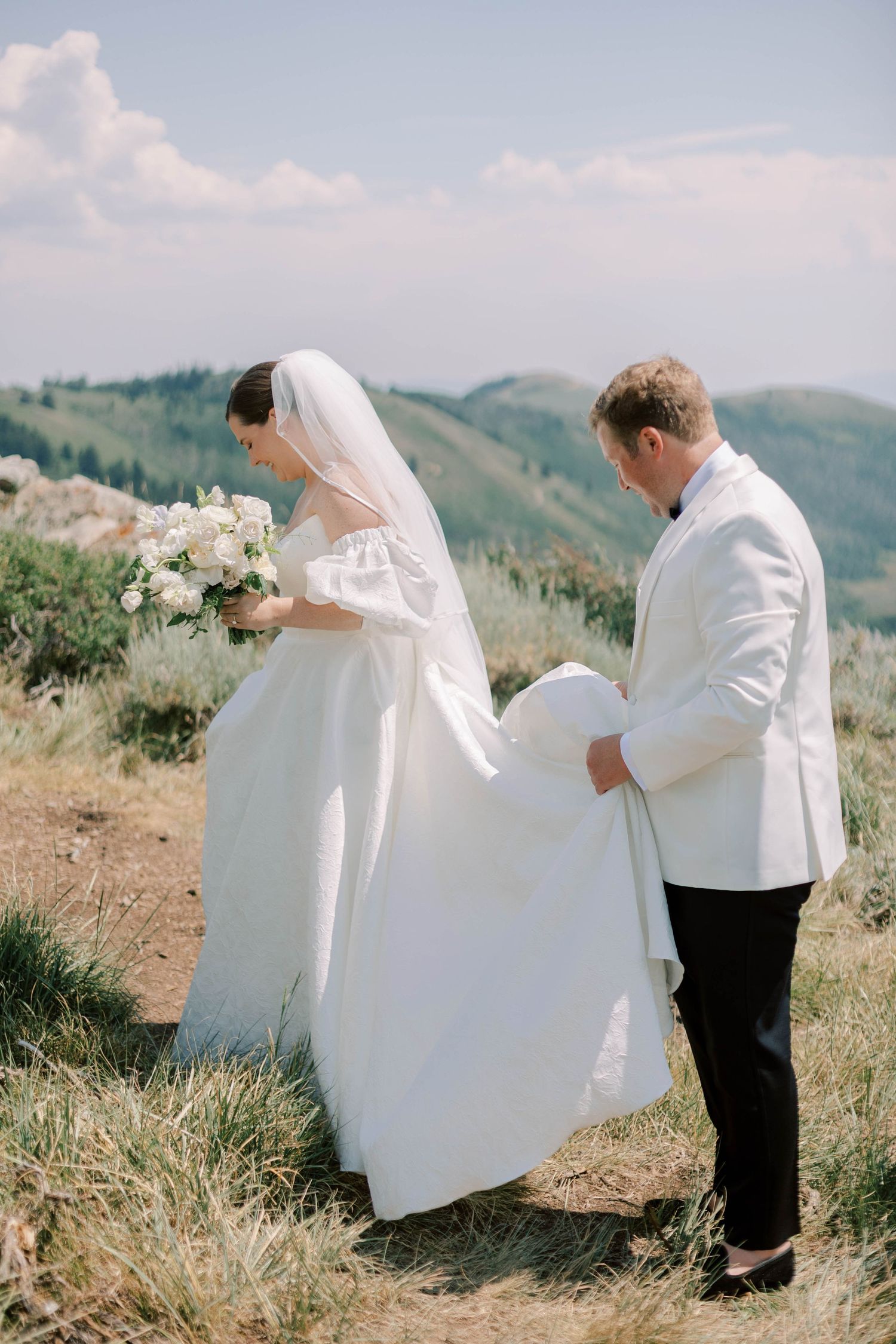 Groom helps bride with dress train on mountain path during wedding photoshoot.
