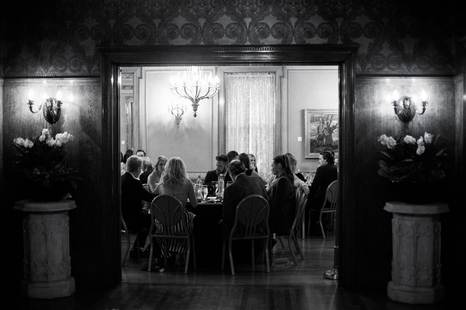 Black and white image of reception dinner through doorway with ornate lighting fixtures.