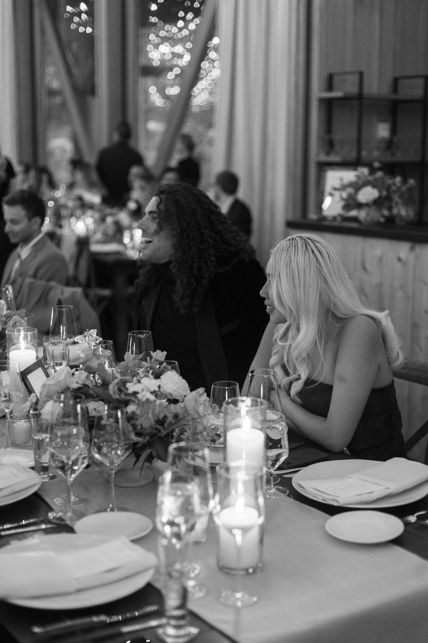 Black and white photo of an elegant dinner party with candlelit table settings and floral centerpieces.