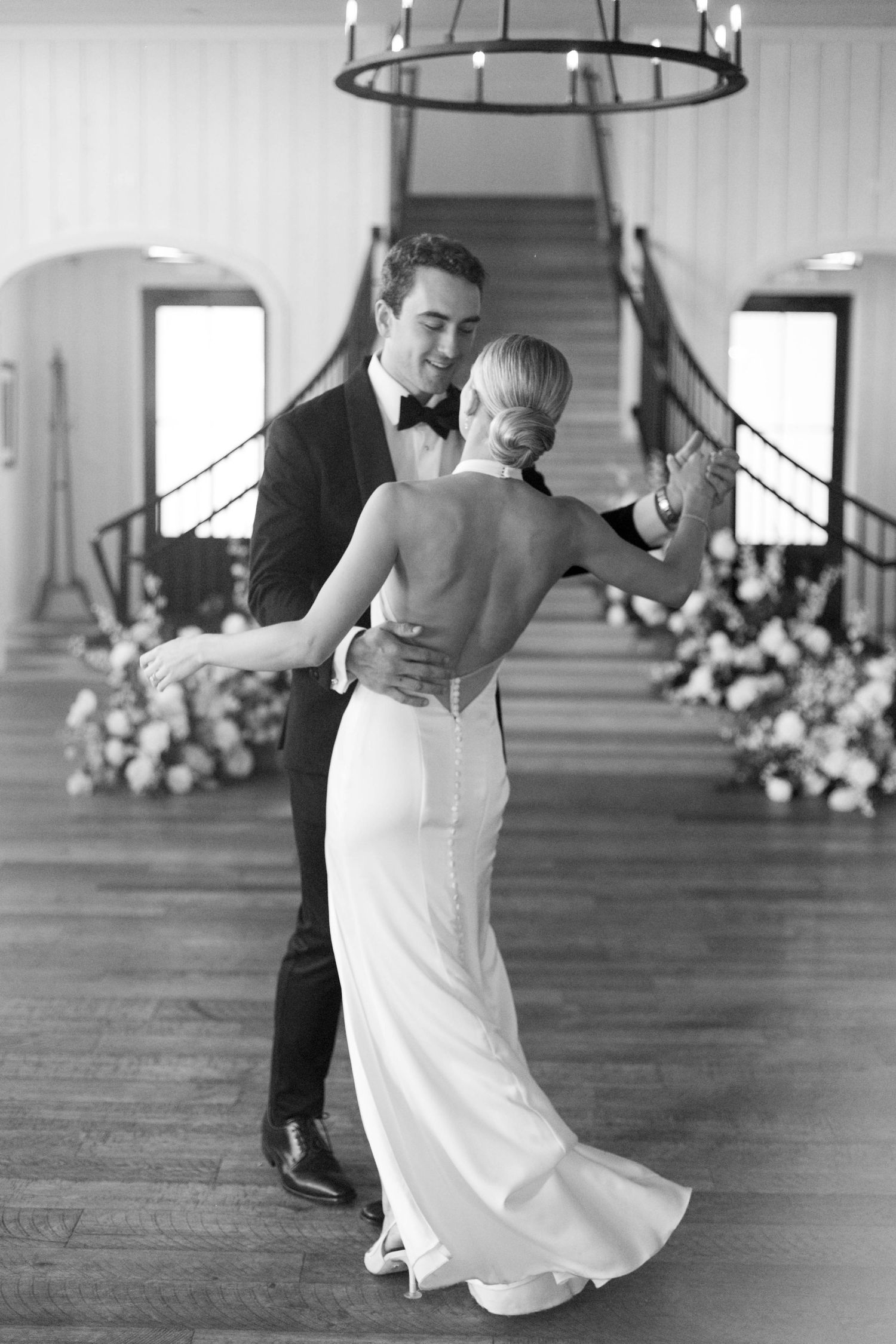 A couple dancing in an elegant venue with a grand staircase and chandelier in black and white.