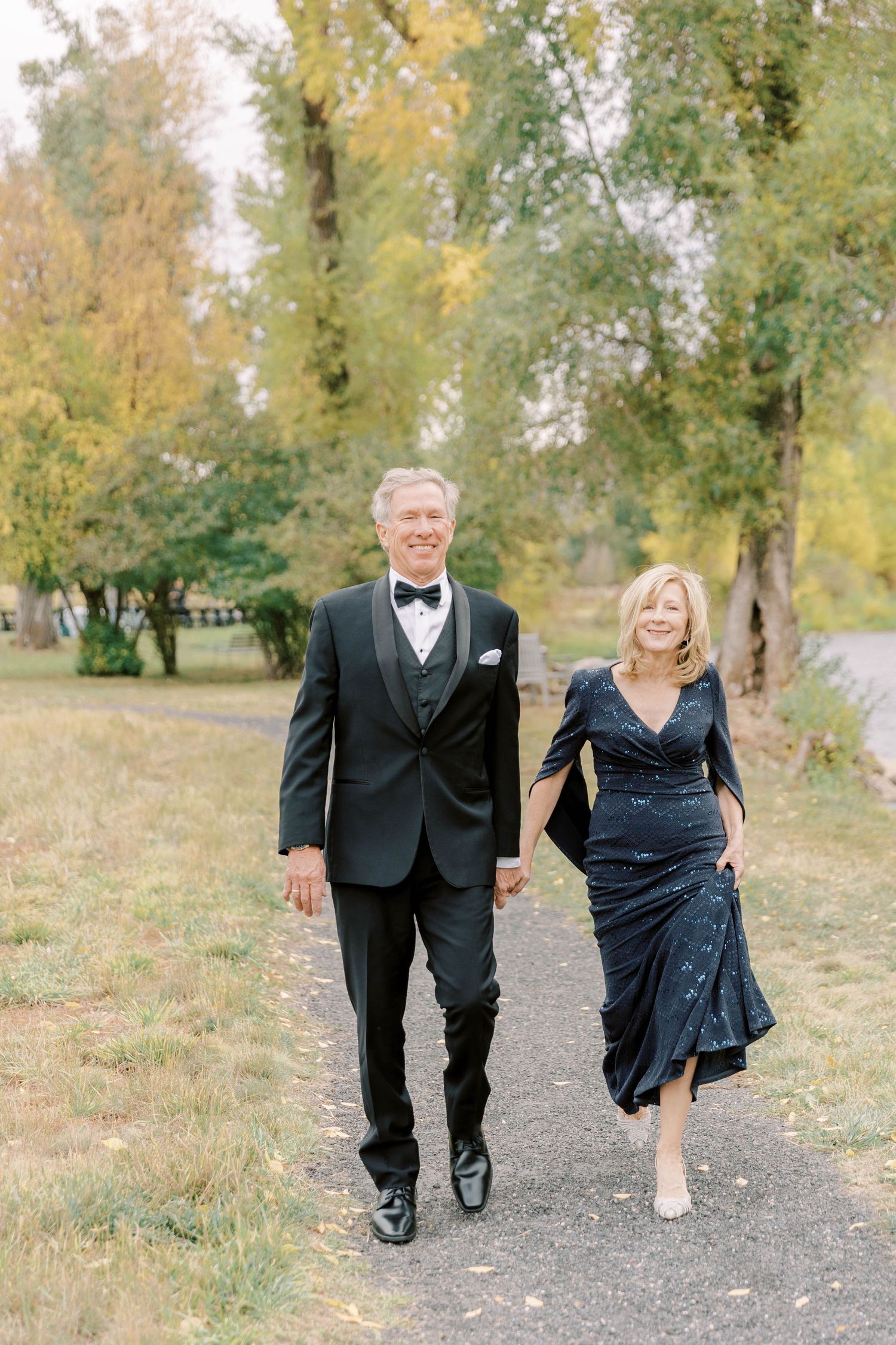 Formal couple in black tie attire walks along autumn path together.