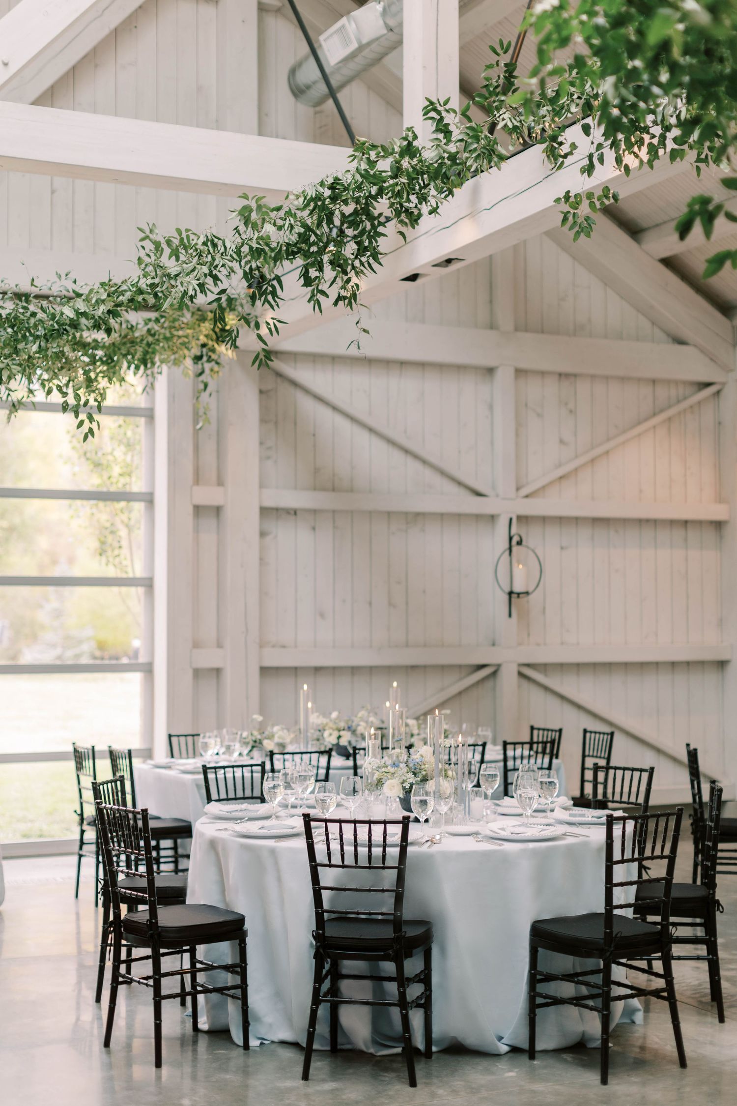 Elegant barn reception space with round tables, black chiavari chairs and hanging greenery.