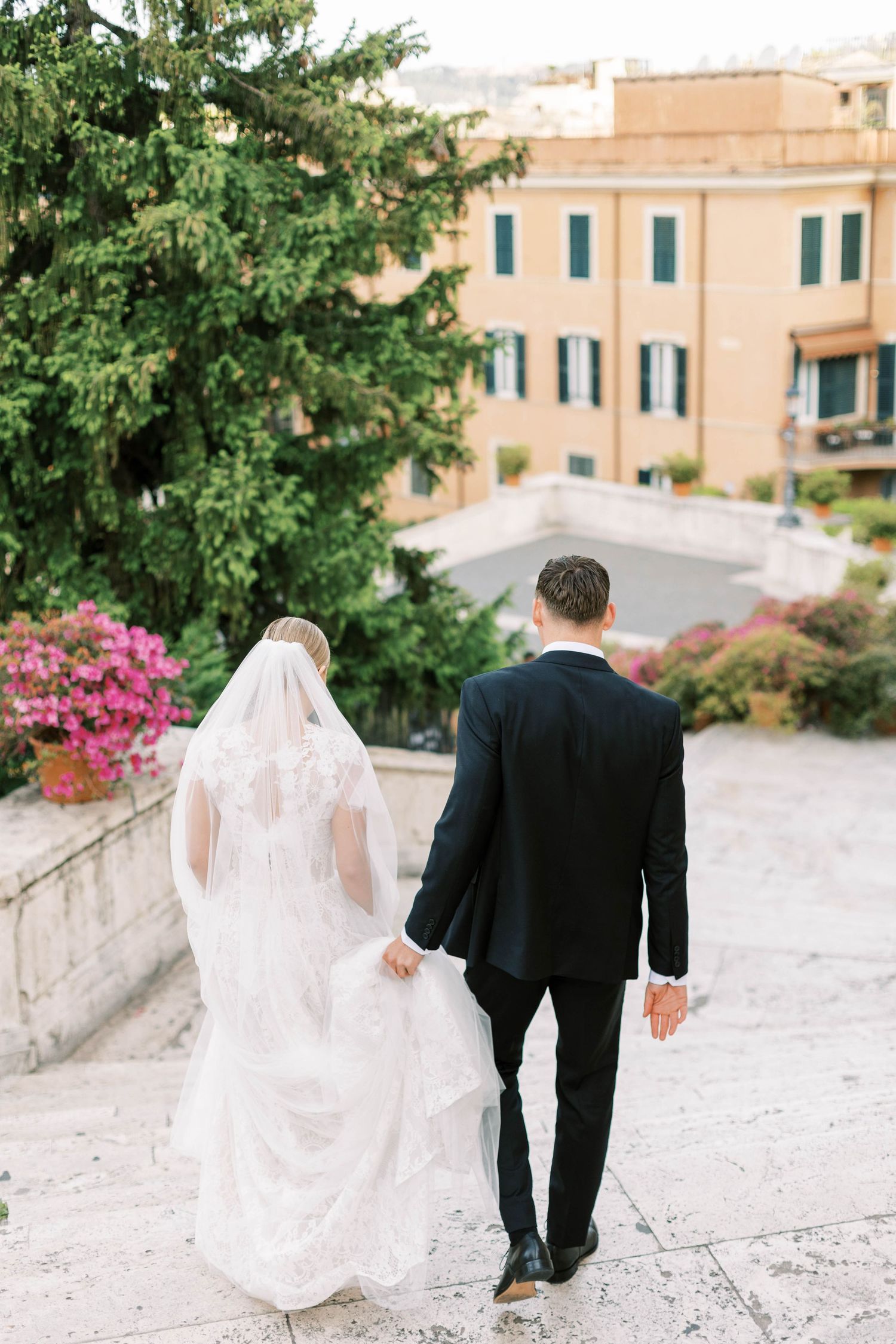 Couple walking hand in hand through a garden path with Italian buildings and pink azaleas in view.