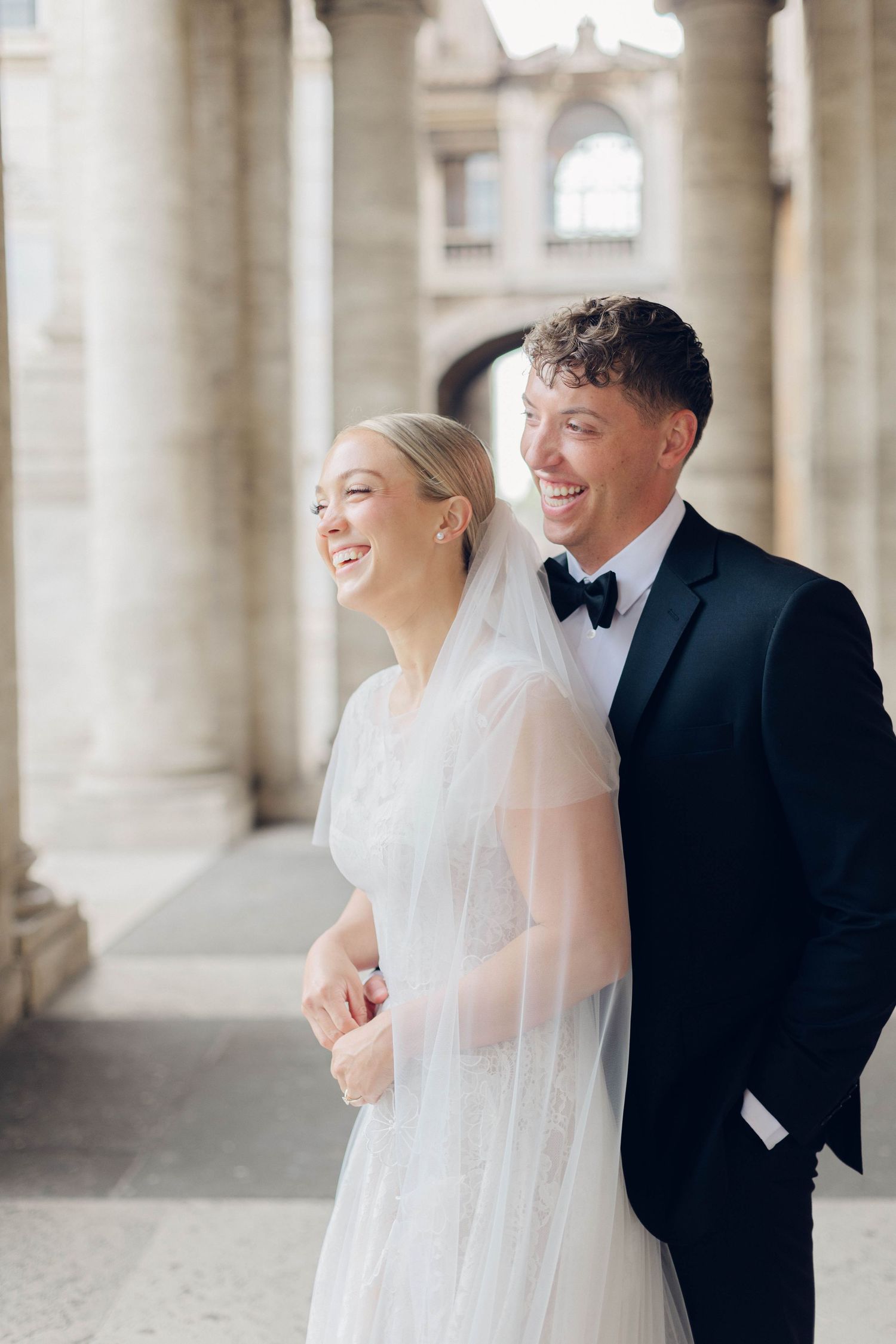 A joyful wedding couple laughs together under ornate stone columns of rome italy.