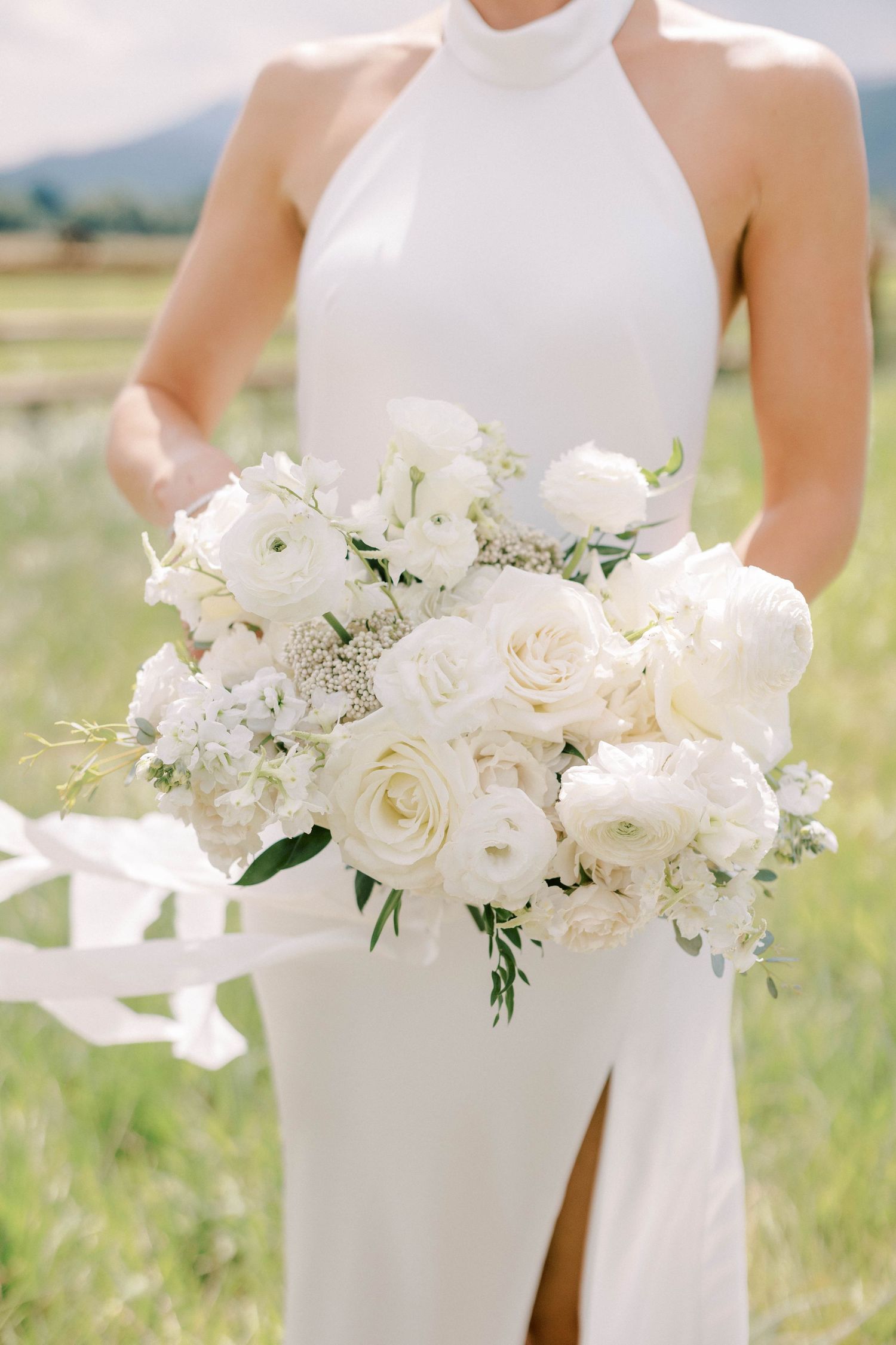Elegant white bridal bouquet with roses and flowing ribbons against mountain landscape.