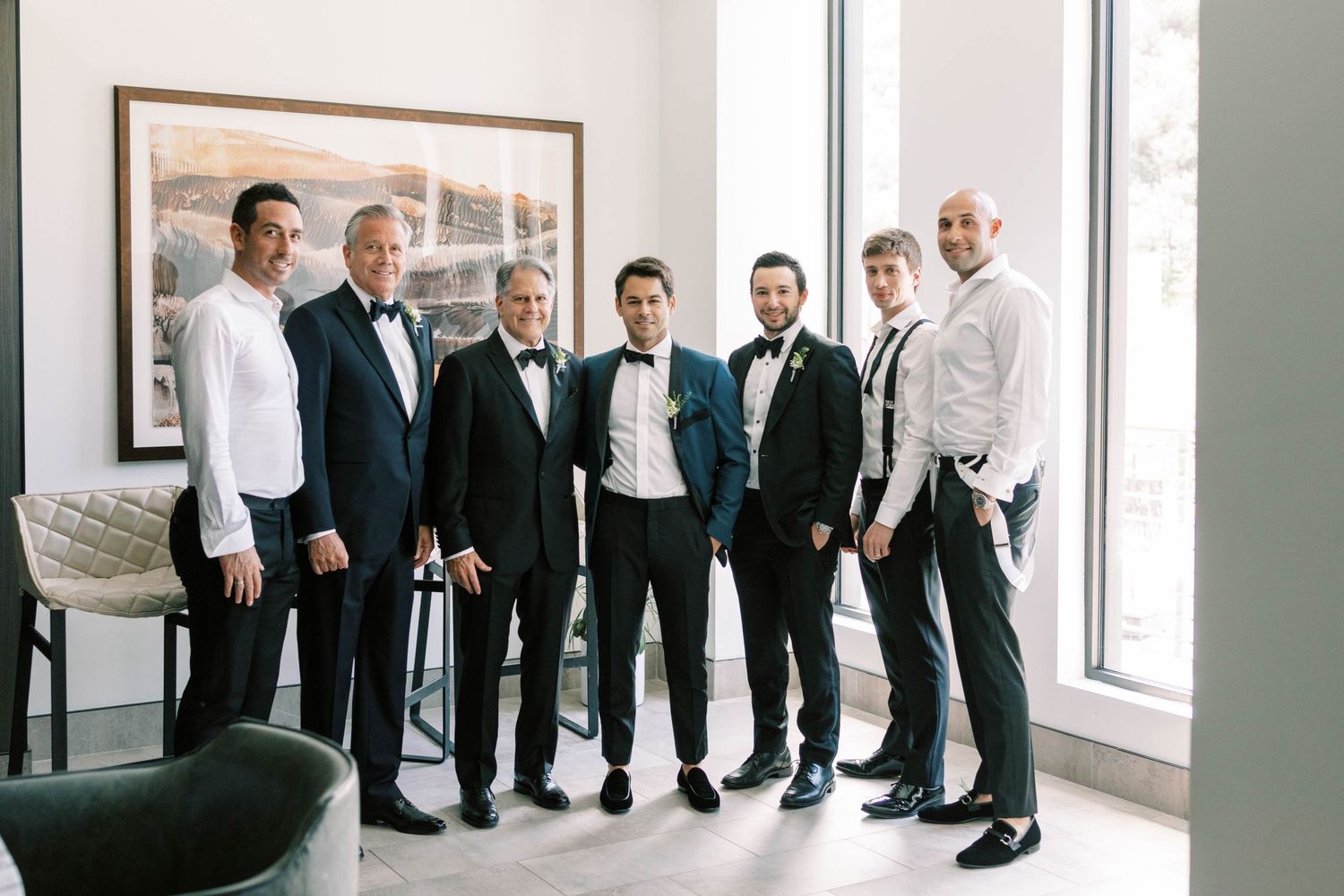 Formal group photo of groomsmen in tuxedos standing by window.