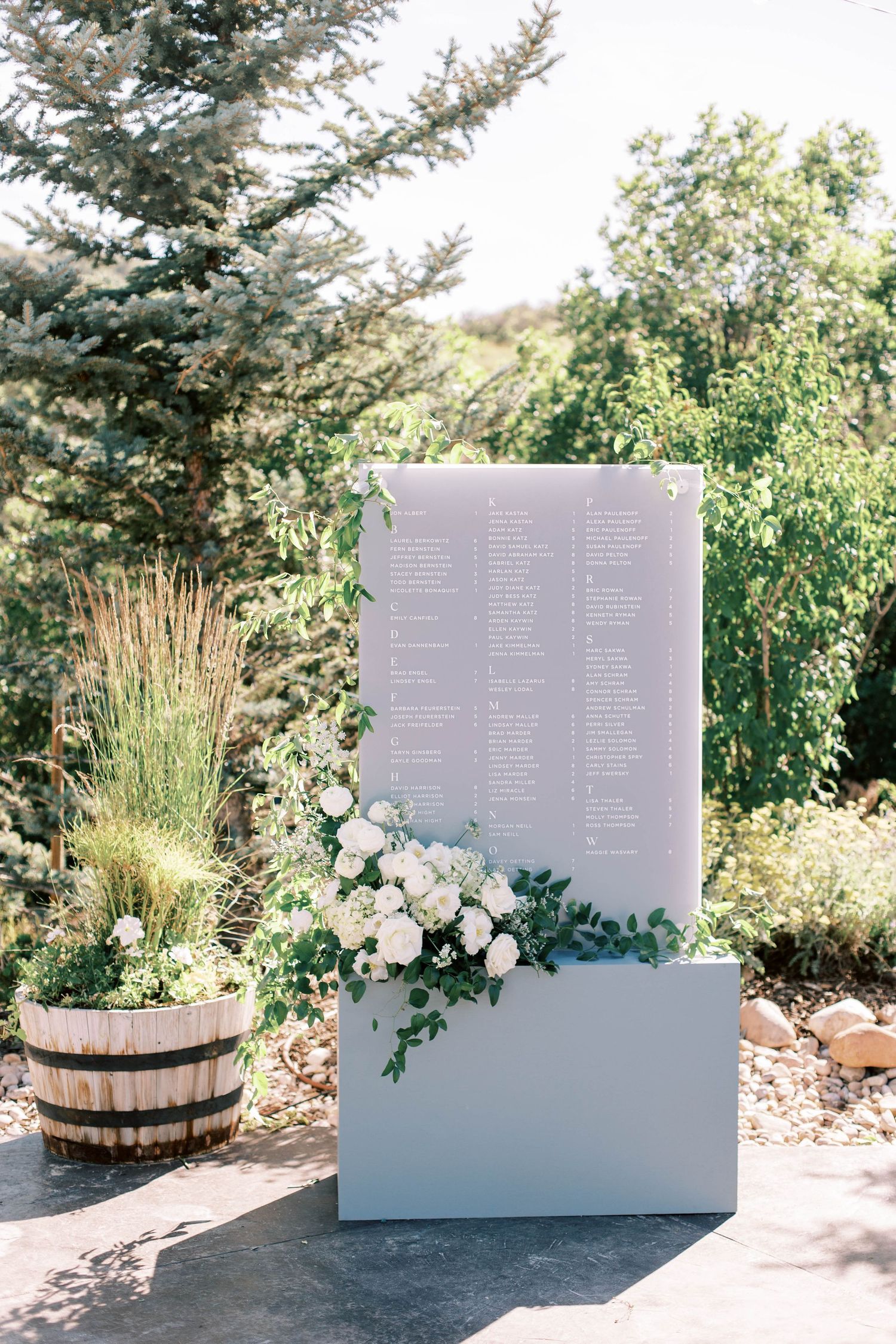 Modern wedding seating chart display with white floral accents against an outdoor garden setting.