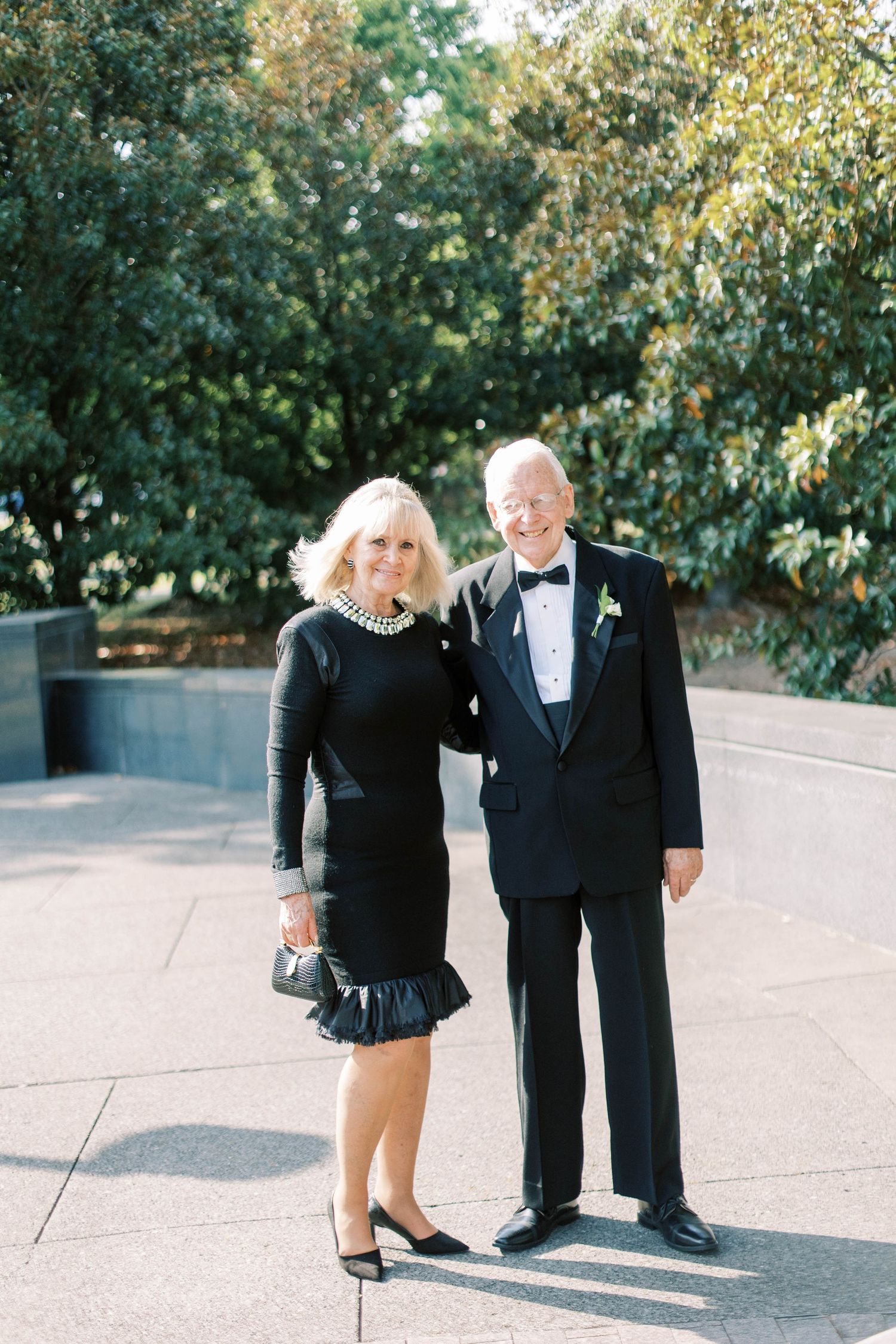 Formal couple in black evening attire pose together at an outdoor venue.