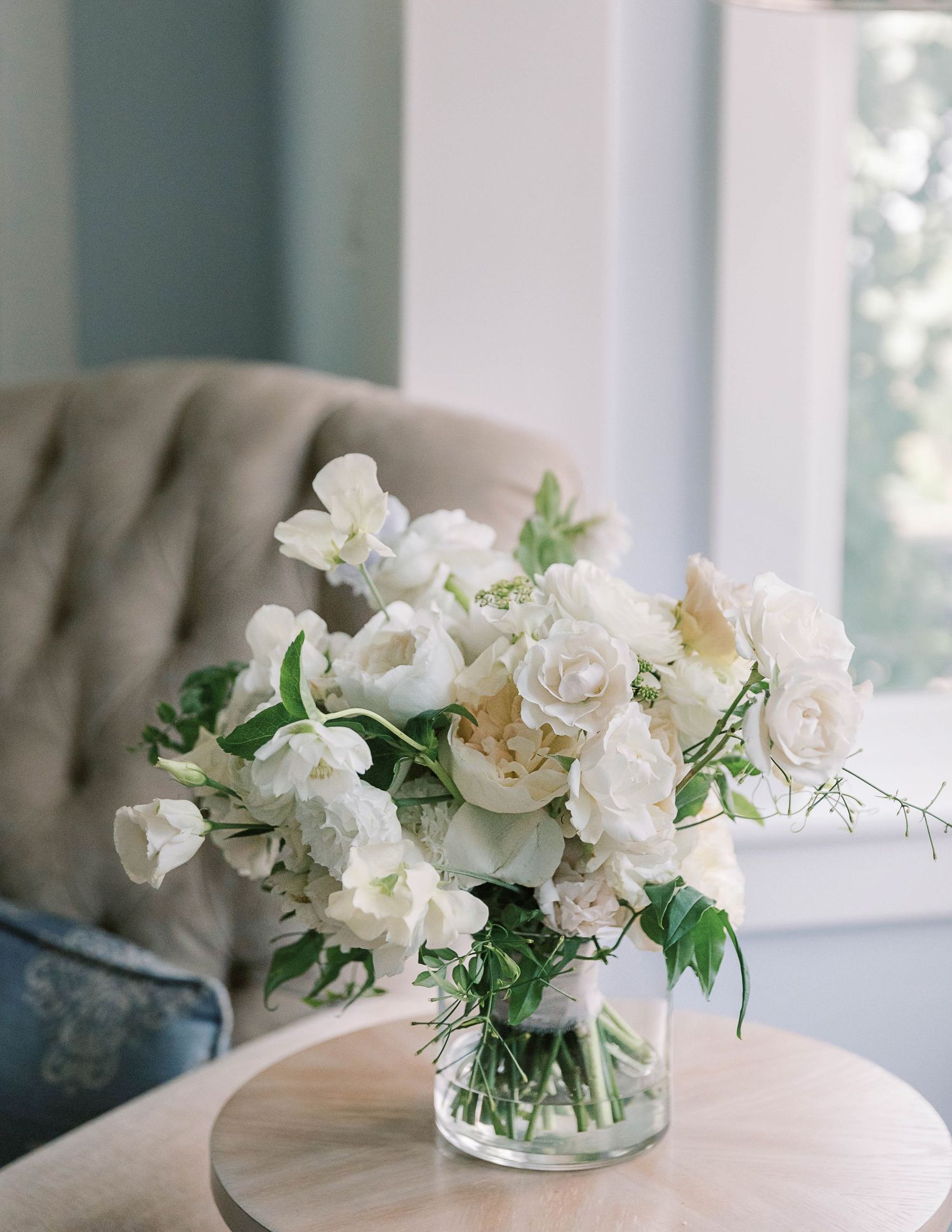 White roses and flowers arranged in a clear glass vase on a side table by a window.