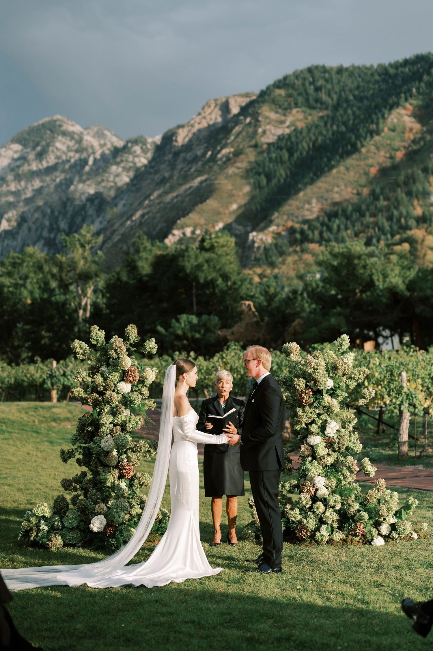 Mountain backdrop frames garden wedding ceremony with lush white floral arch installation.