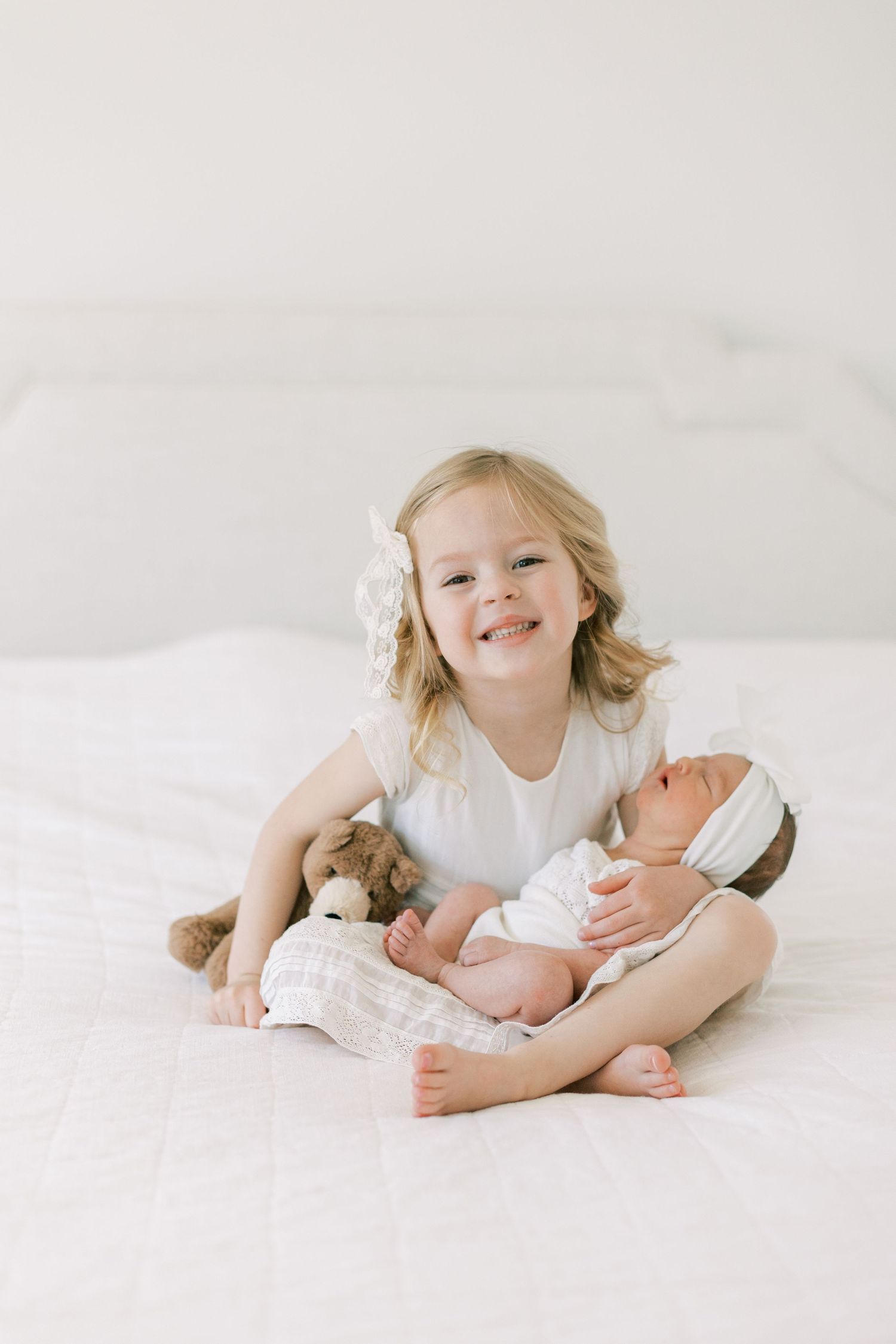 Young girl holding newborn sibling while sitting on white bedding with stuffed toy.
