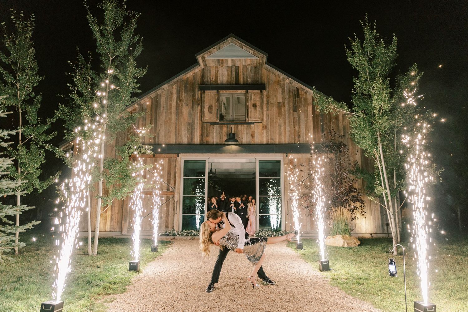 Romantic couple pose in front of rustic barn venue with sparkling light fountains illuminating the path.