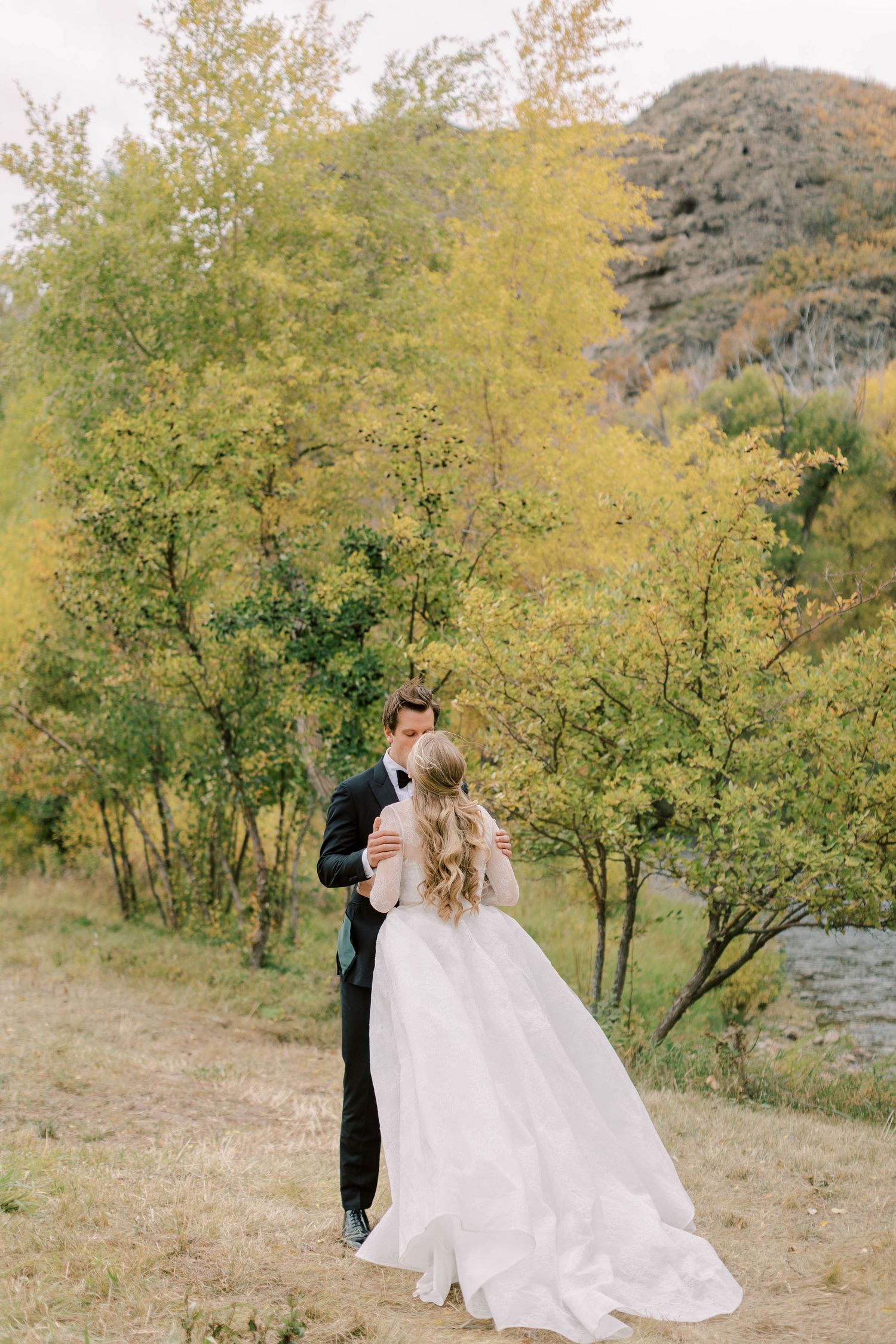 A romantic wedding couple embraces among yellow fall foliage near a mountain stream.