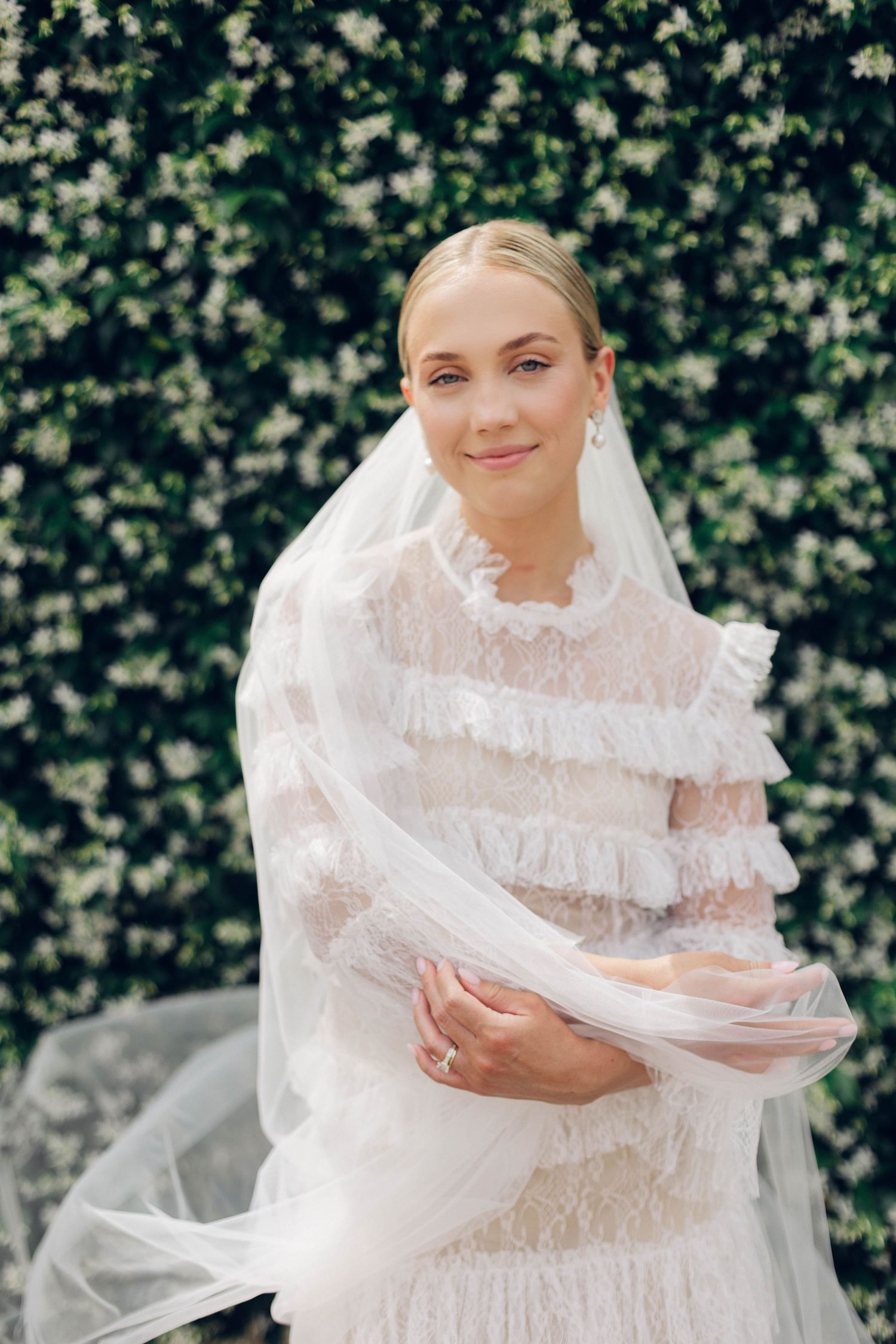 A bride in a tiered lace wedding dress and veil poses against flowering greenery.