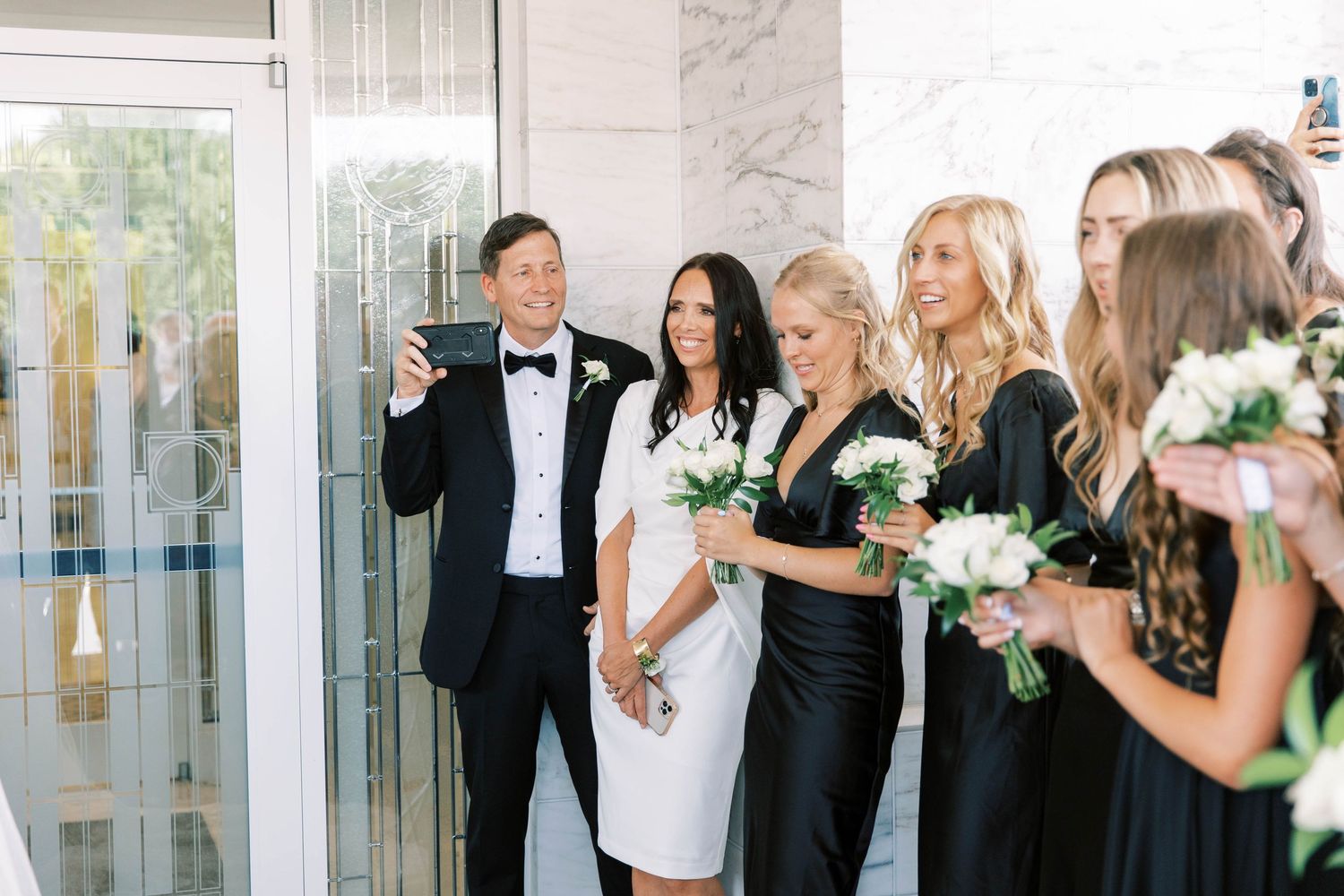 Wedding party poses with bouquets by glass doors in formal attire.
